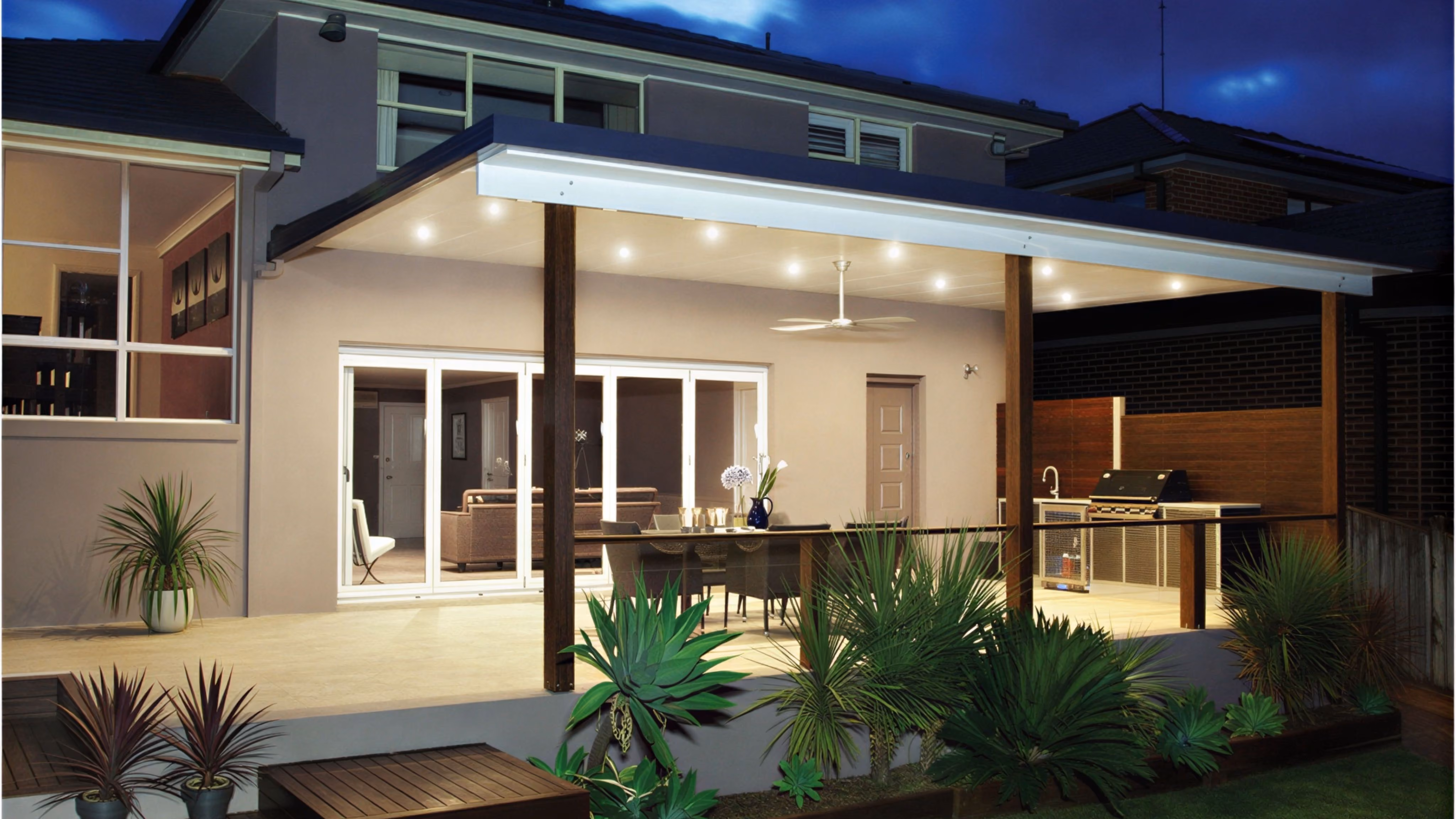 Backyard patio at dusk with covered dining area, ceiling fan, built-in grill, and surrounding plants.