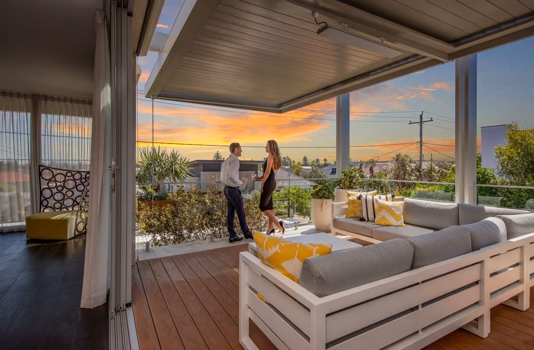 Man and woman talking on a modern outdoor balcony with grey and yellow sofas at sunset.