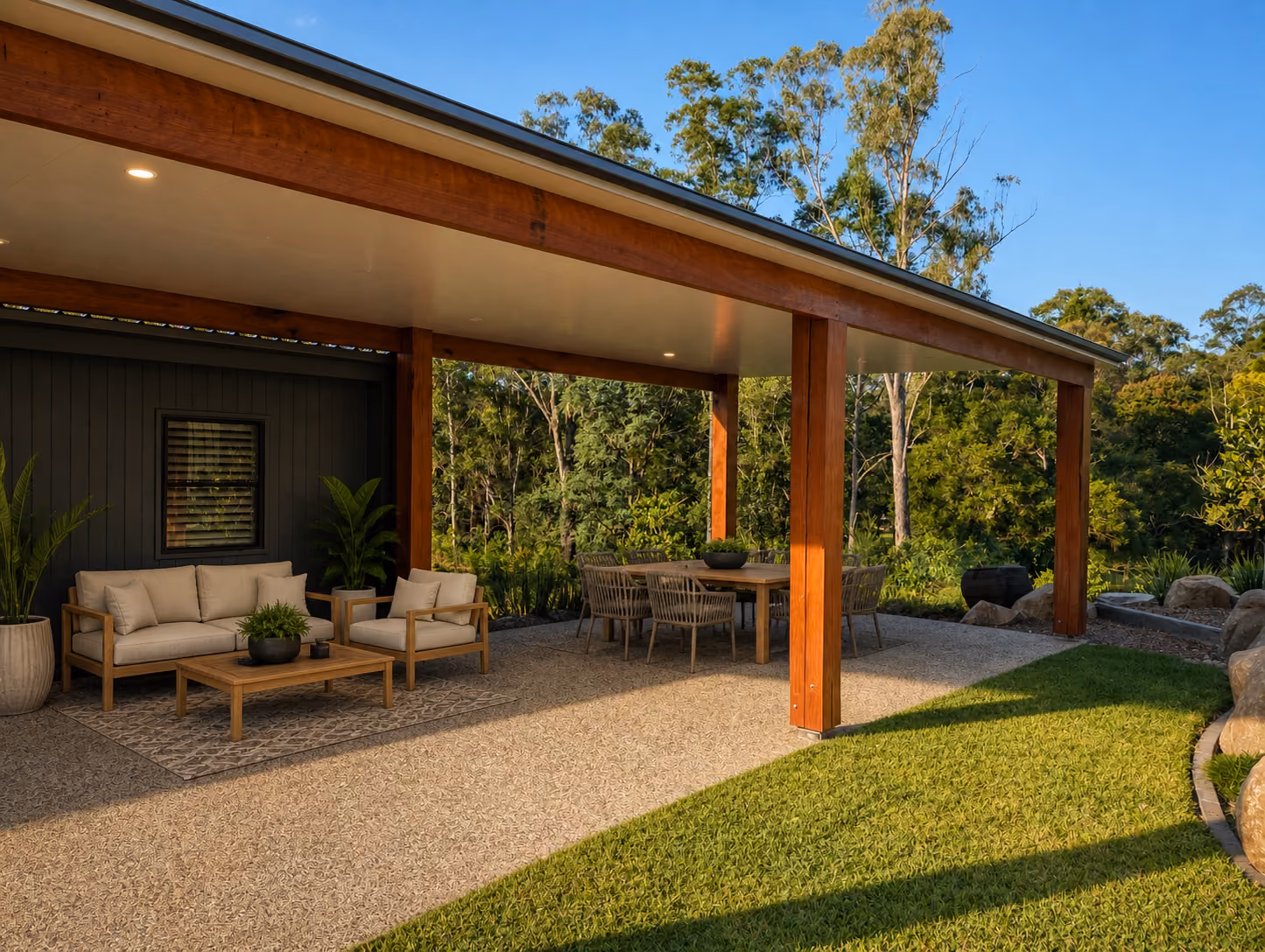 Covered patio with wooden beams, outdoor sofa set, dining table, and surrounding greenery under blue sky.