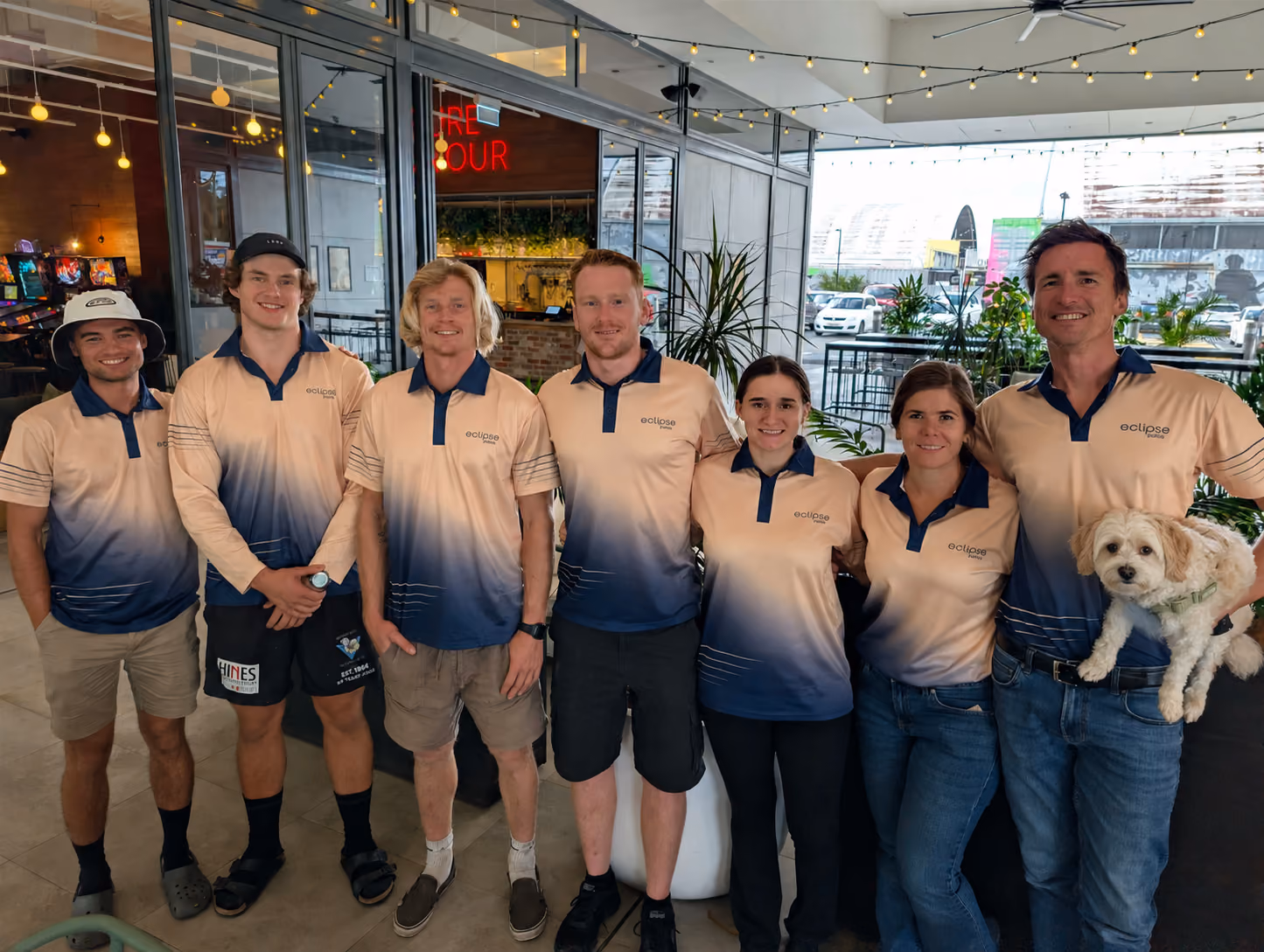Seven people wearing matching eclipse polos stand inside a café, with one man holding a small dog.