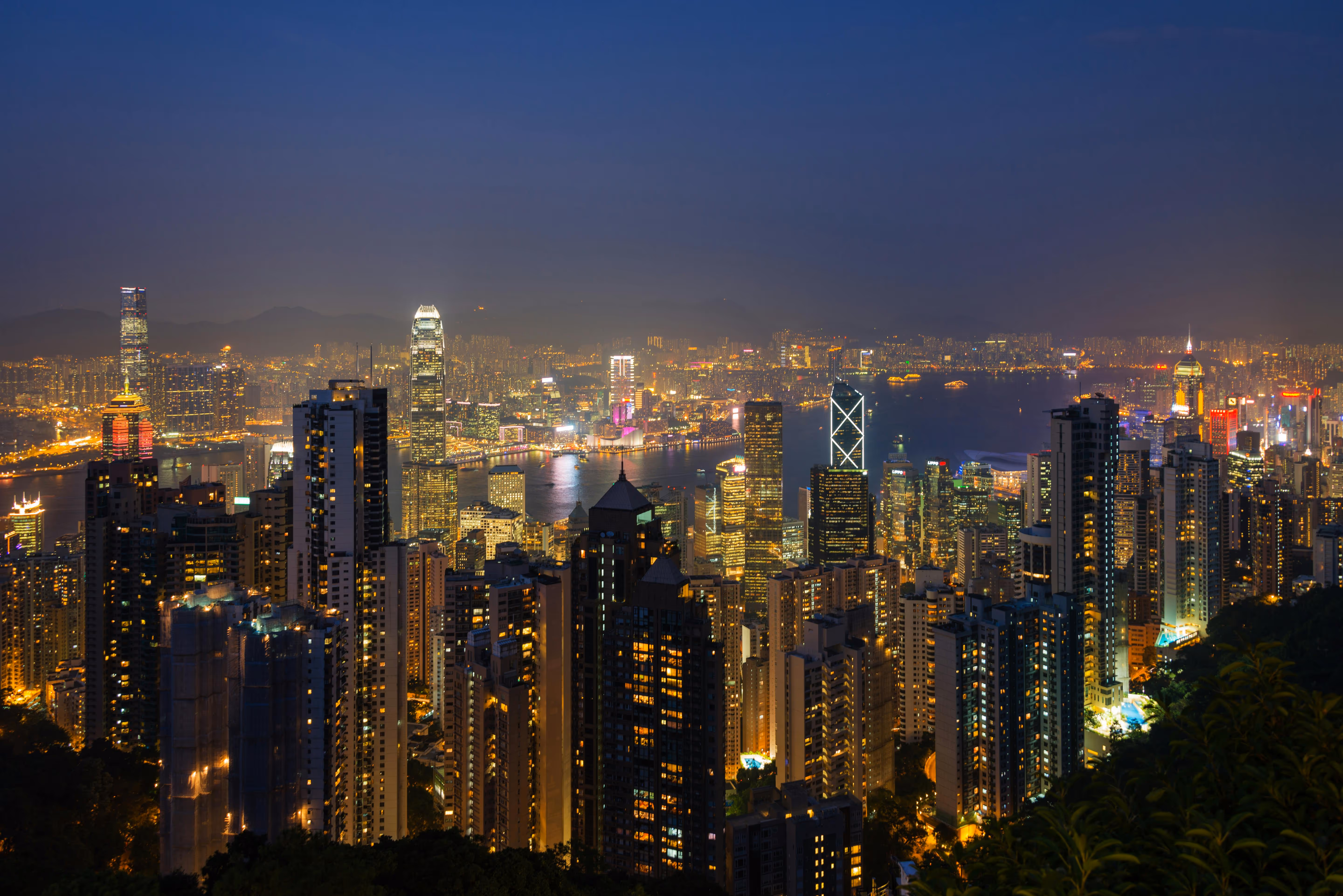 Nighttime cityscape of a densely built urban area with illuminated skyscrapers and a harbor reflecting city lights.