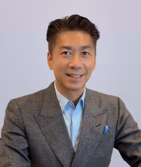 Smiling man with dark hair wearing a grey blazer and light blue shirt against a plain background.