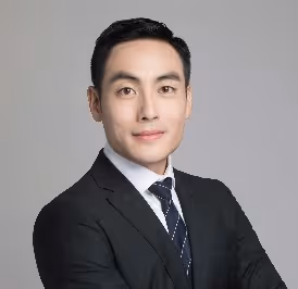 Man in a black suit with a white shirt and dotted dark tie, posing against a plain gray background.