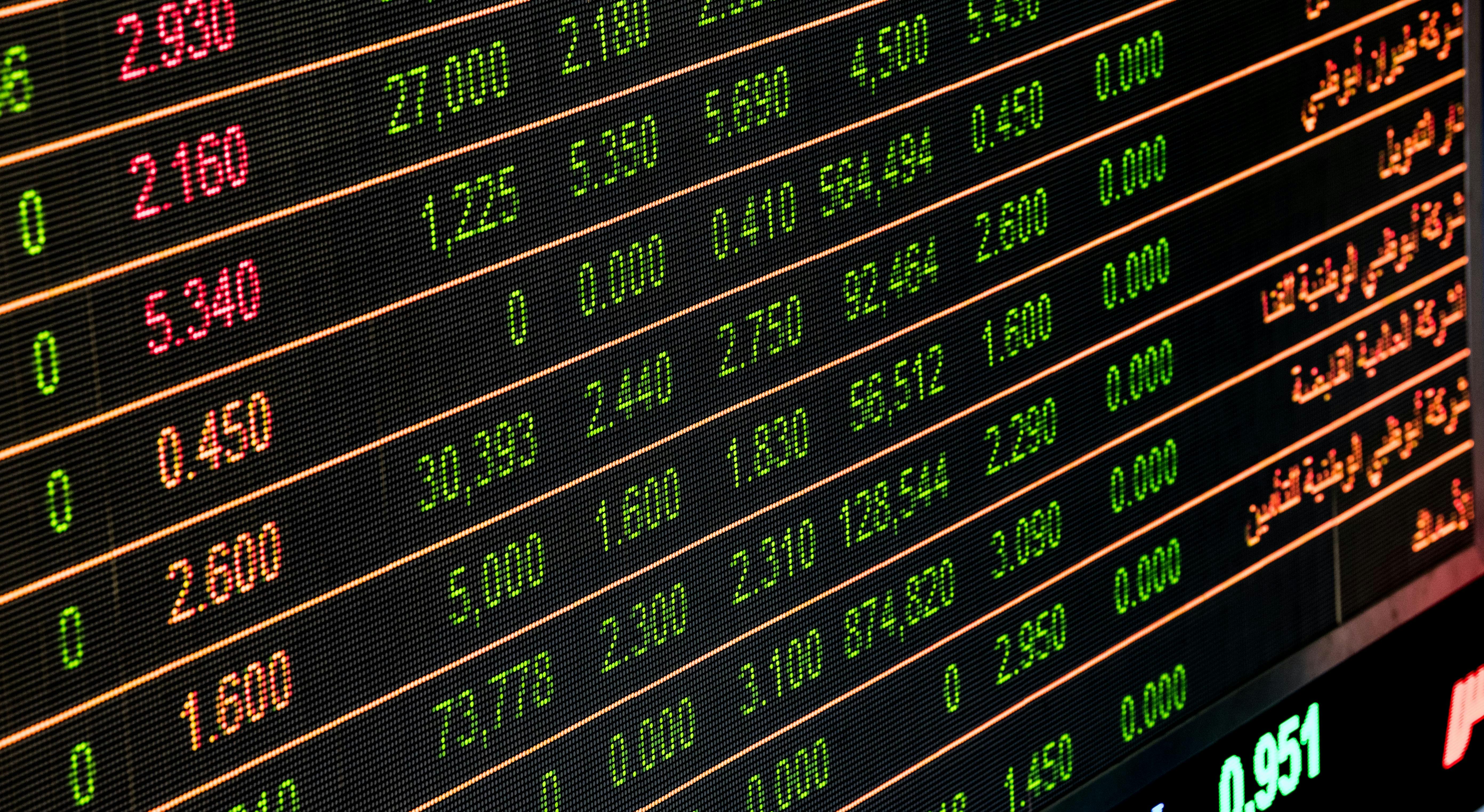 Electronic stock market display board showing financial numbers in green, red, and orange on a black background with some Arabic text on the right.