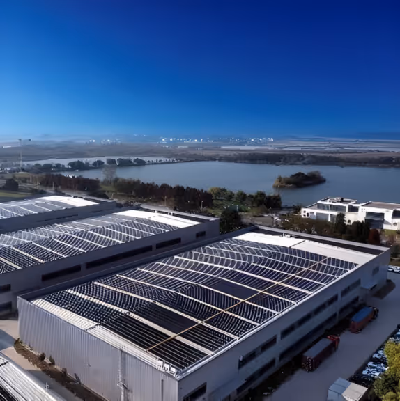 Aerial view of industrial buildings with solar panels on roofs near a large river under a clear blue sky.