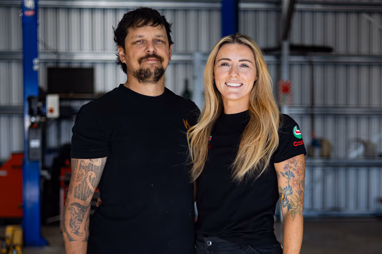 Smiling man and woman standing side by side in a garage or workshop with metal walls.
