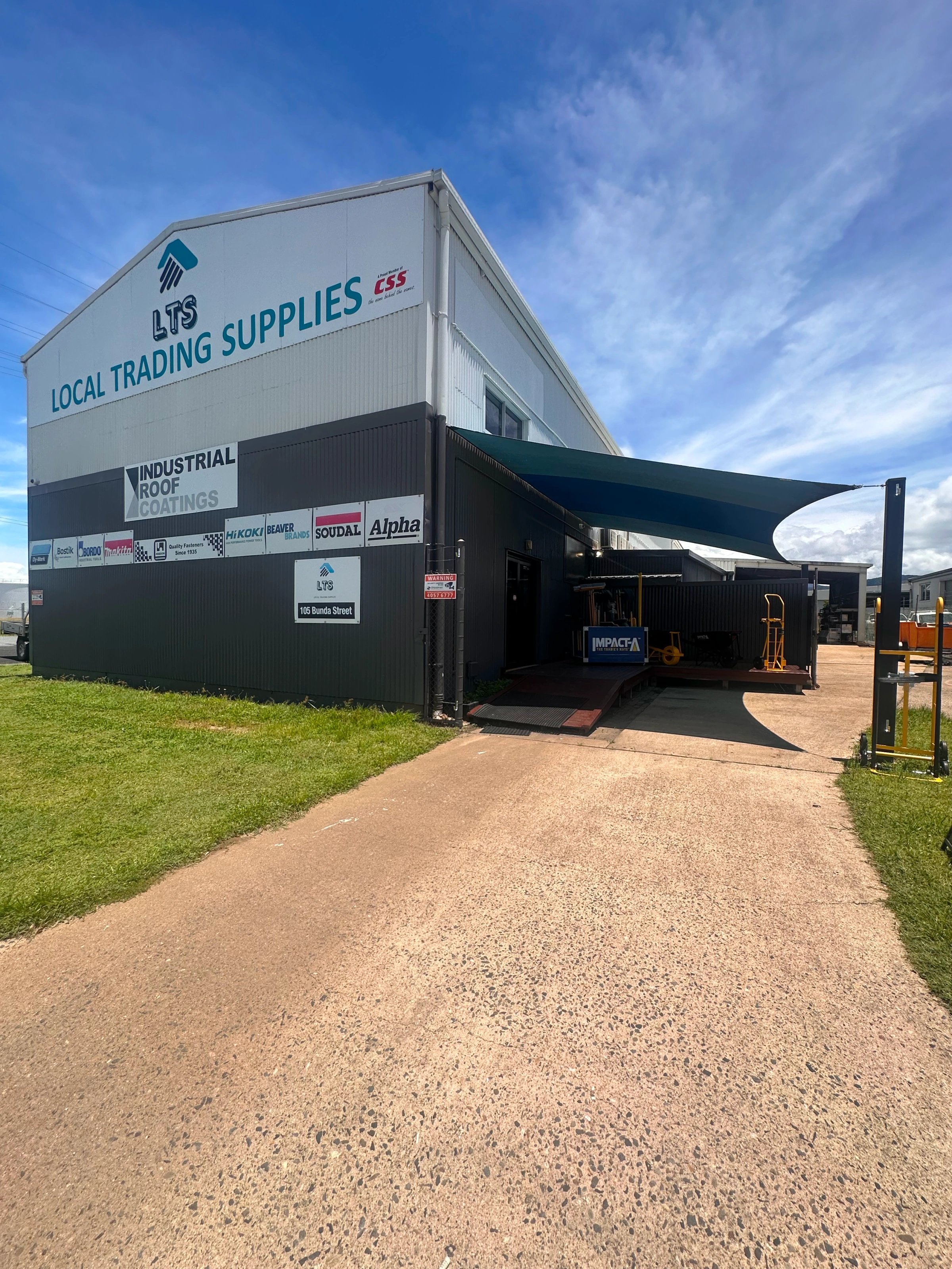 Commercial building labeled Local Trading Supplies with multiple industrial roof coating brand signs under a blue sky.