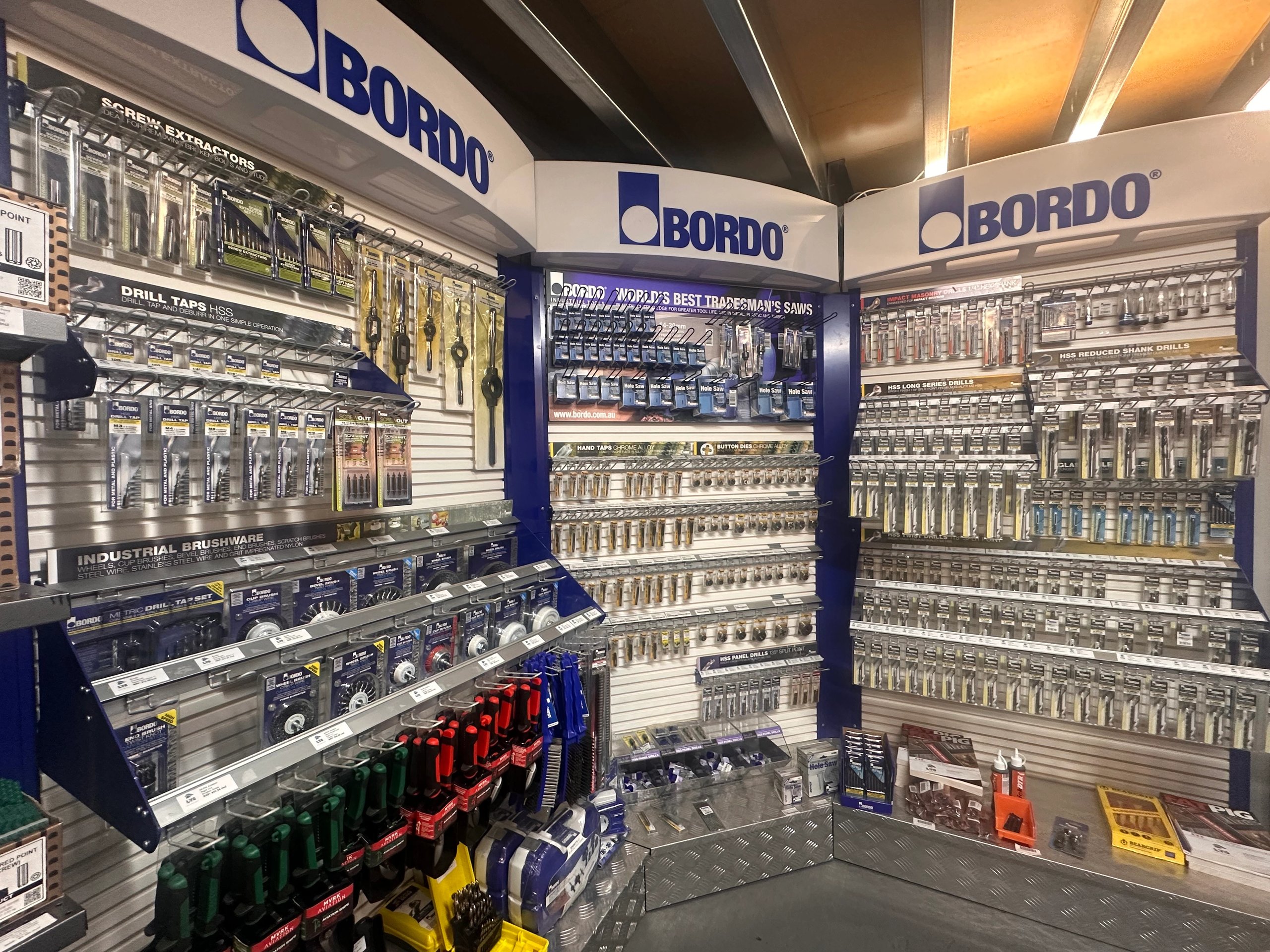 Wall display of various Bordo branded drill taps, screw extractors, industrial brushes, hole saws, and drill bits organized in packaging in a hardware store.