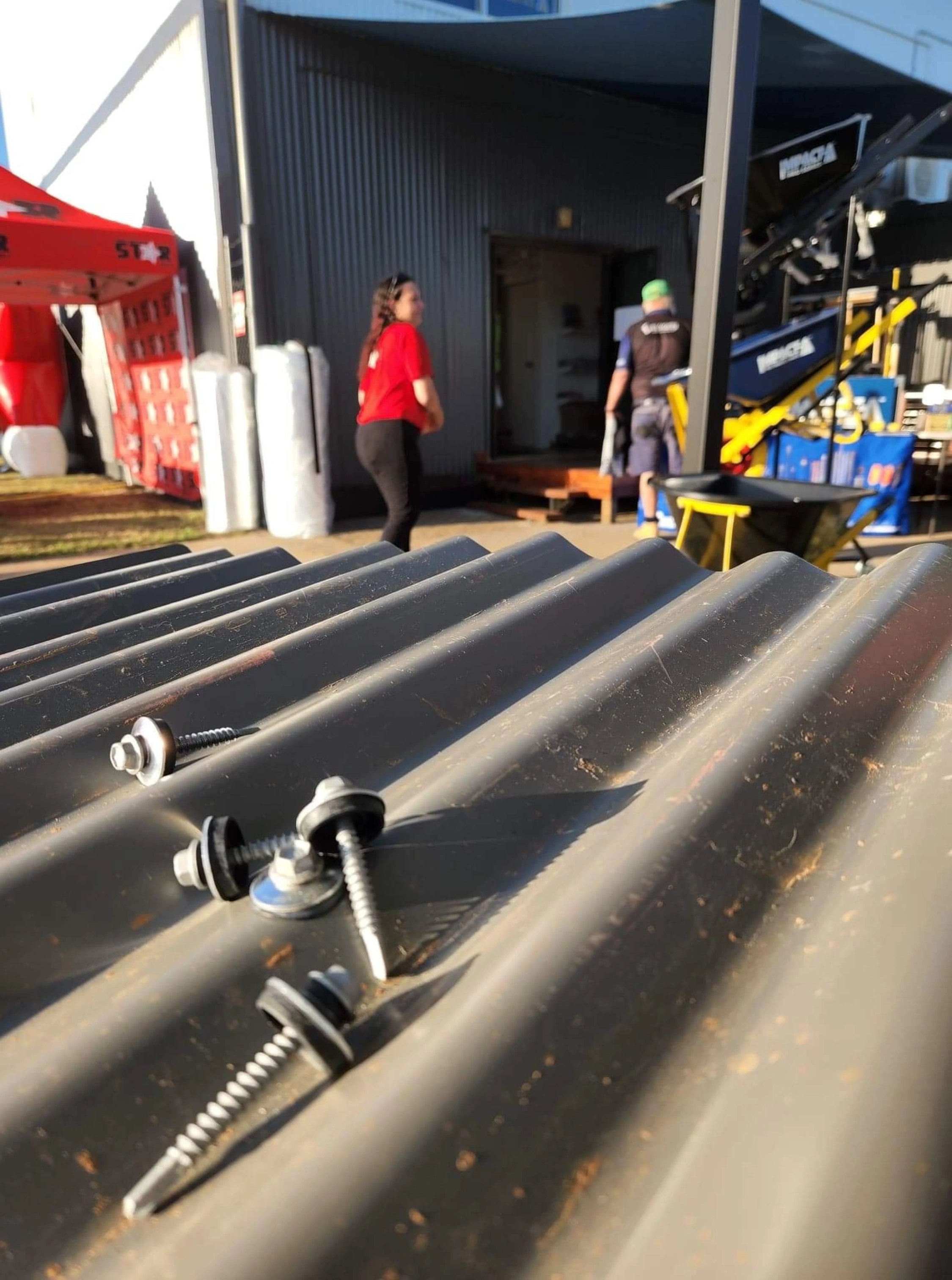 Close-up of several screws on a corrugated metal surface with people and industrial equipment in the background.