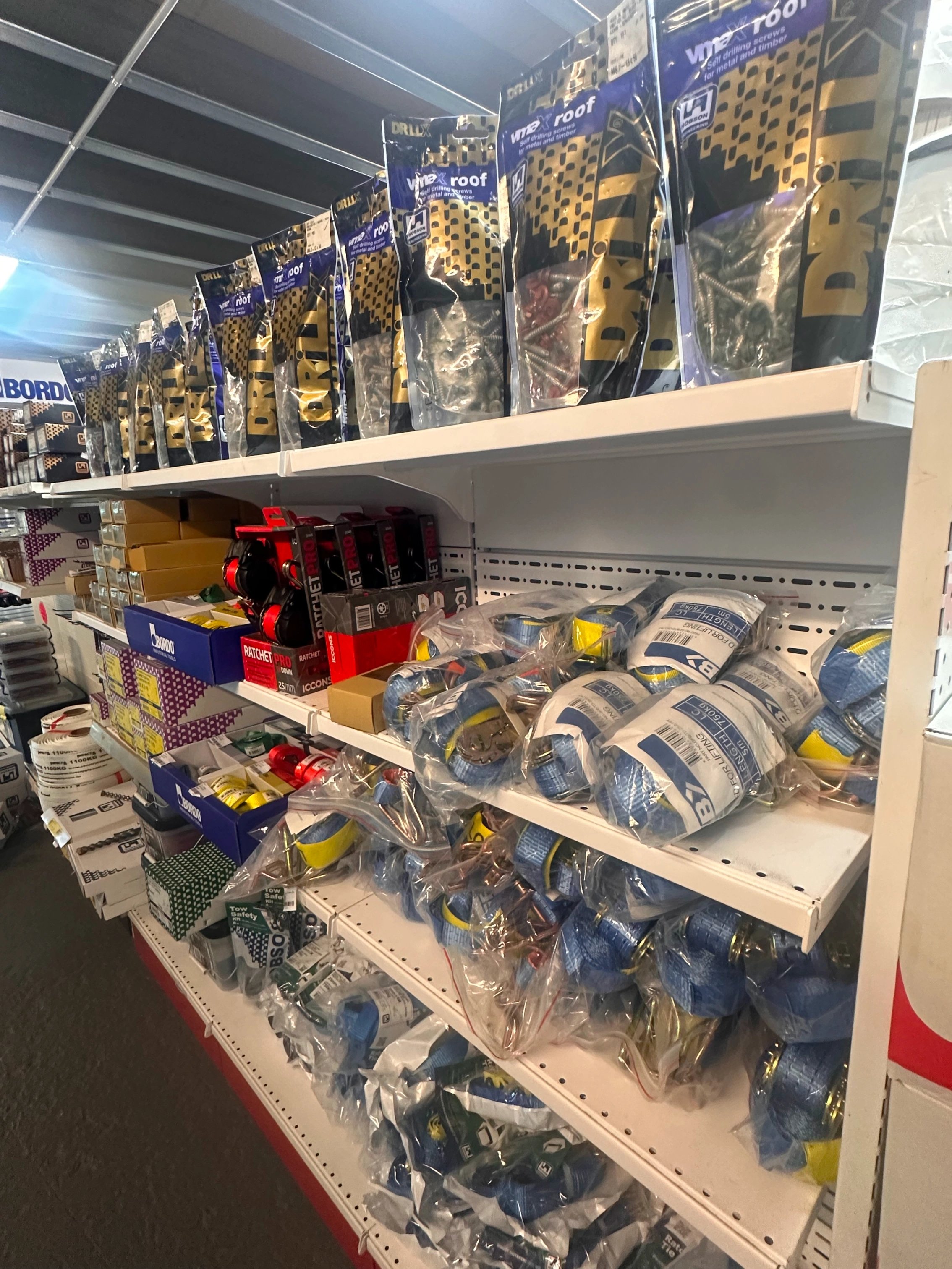 Shelves stocked with assorted hardware supplies including bags of screws, ratchet straps, and other packaged tools in a store aisle.