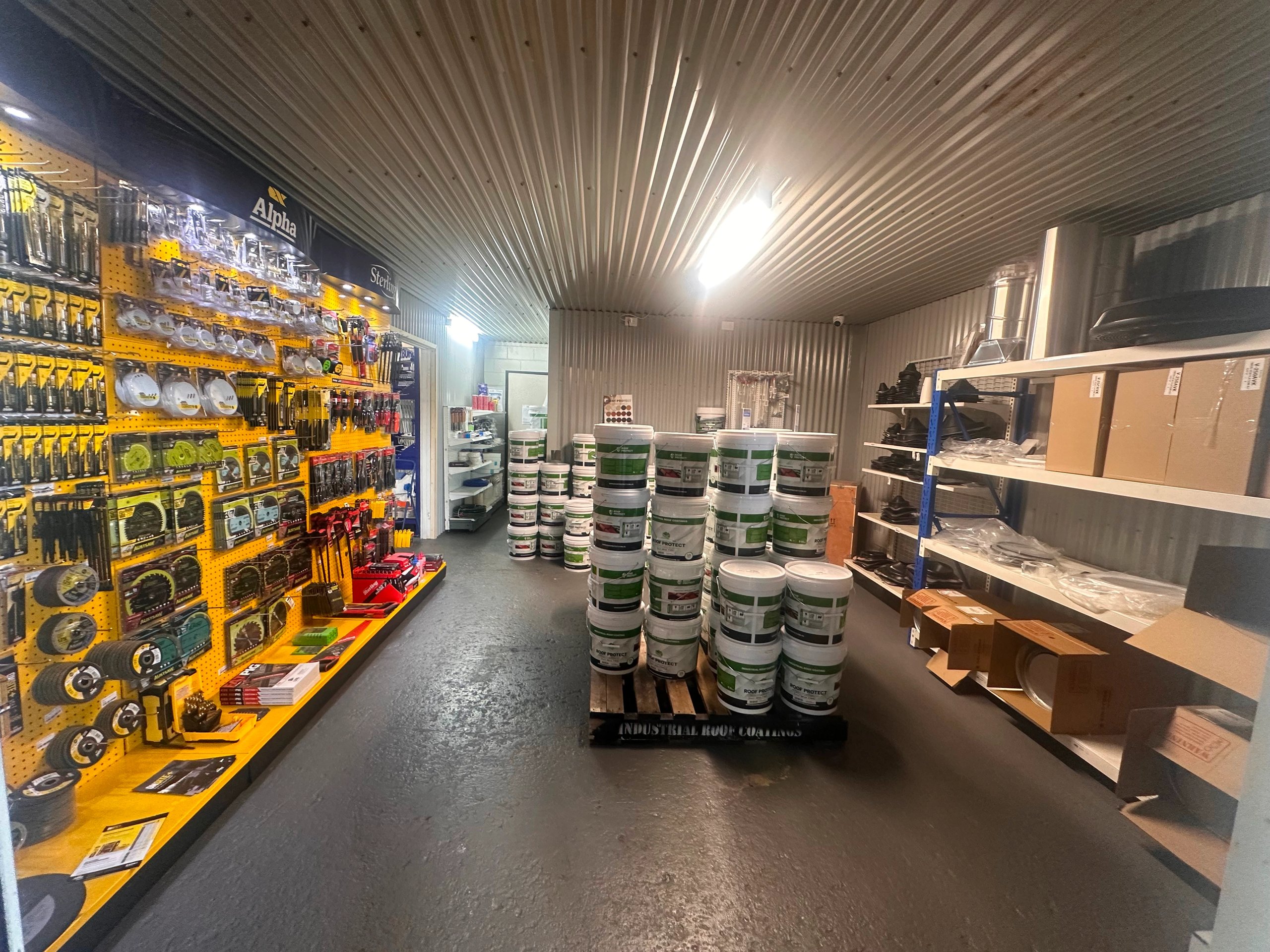 Interior of a hardware store with shelves displaying tools on the left and stacks of white buckets labeled 'Roof Protect' in the center.
