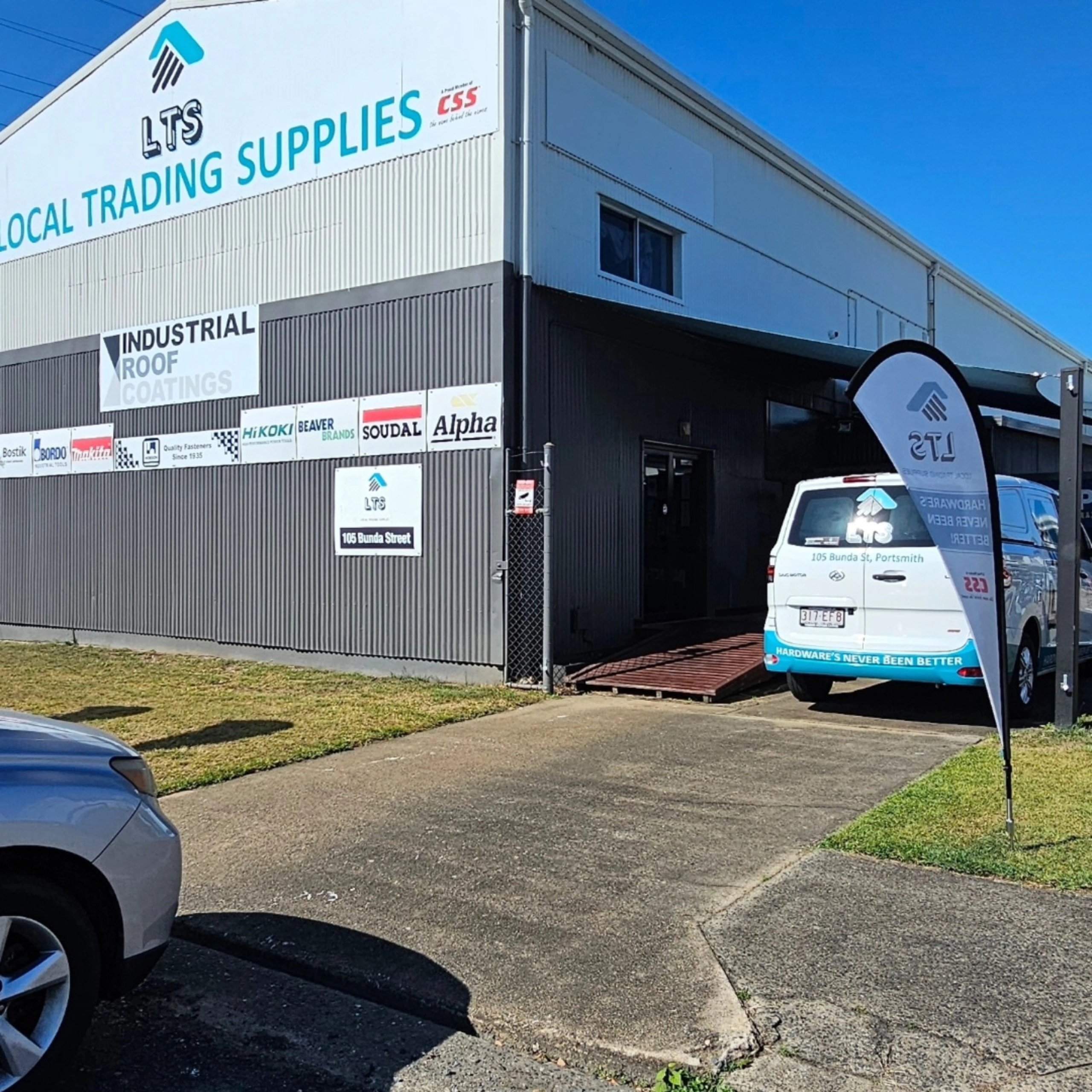 Exterior of a Local Trading Supplies building with signage, a parked company van, and a flag advertising hardware supplies.