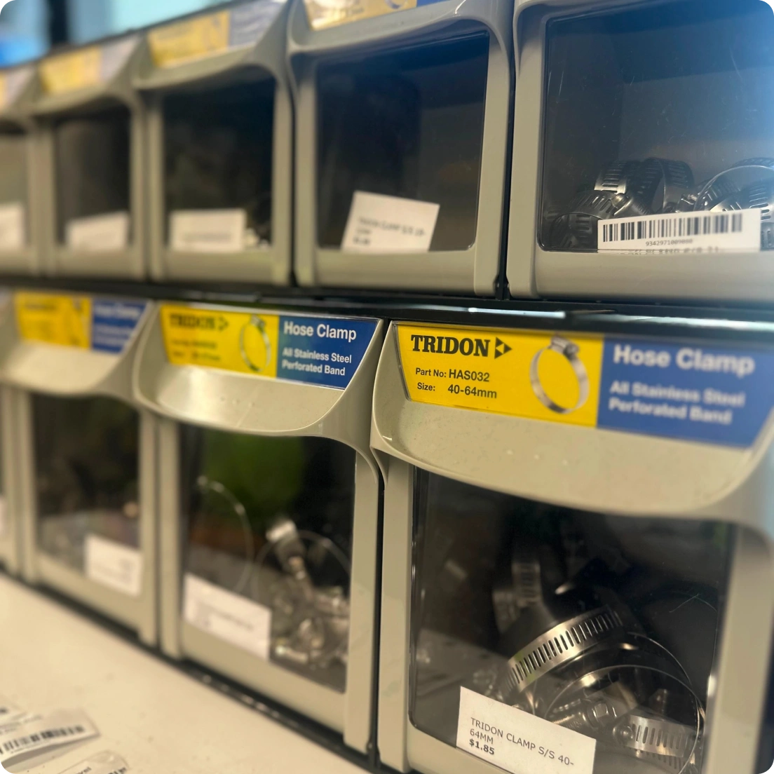 Gray plastic storage bins holding stainless steel hose clamps with yellow and blue TRIDON labels showing part number and size.