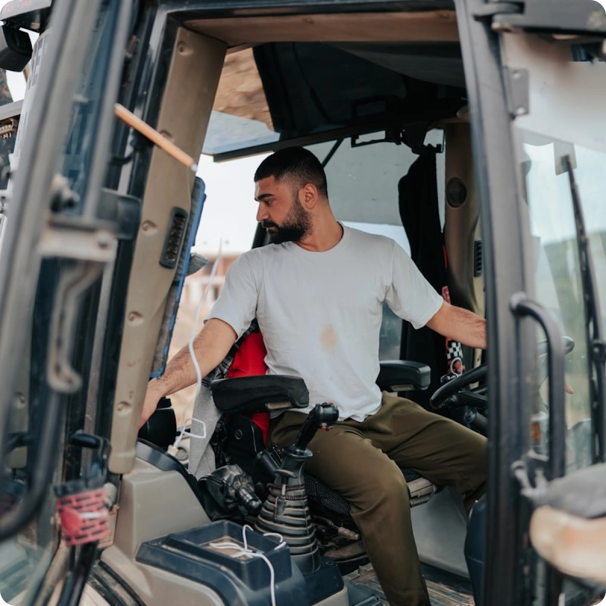 Man in a white t-shirt and olive pants operating controls inside a construction vehicle cab.