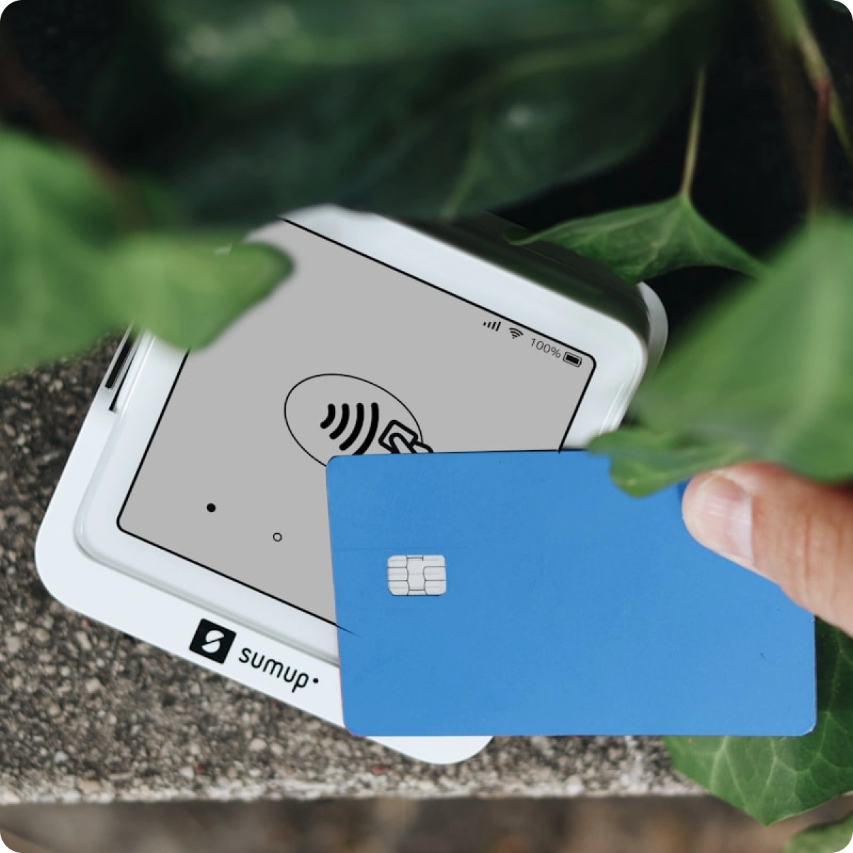 Hand holding a blue credit card near a white SumUp contactless card reader surrounded by green leaves.