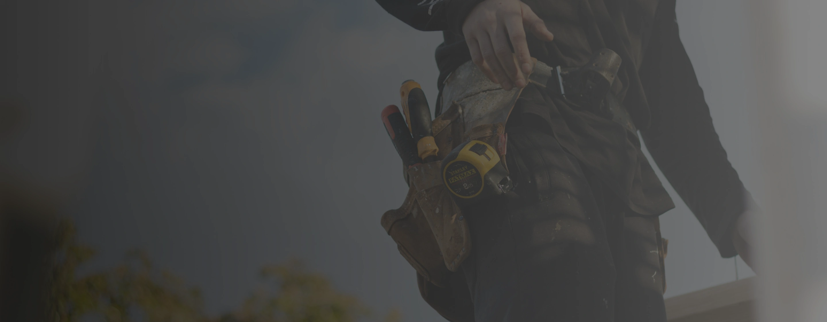 Close-up of a person wearing a tool belt with a tape measure and screwdrivers outdoors.