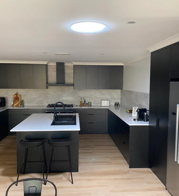 Modern kitchen with black cabinets, white countertops, central island with sink and two black stools, bright ceiling light, and light wood flooring.