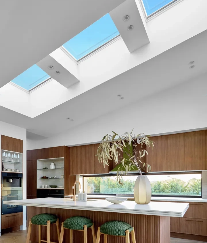 Modern kitchen with wooden cabinetry, a white island countertop with three green stools, a tall vase with leaves, and large skylights letting in natural light.