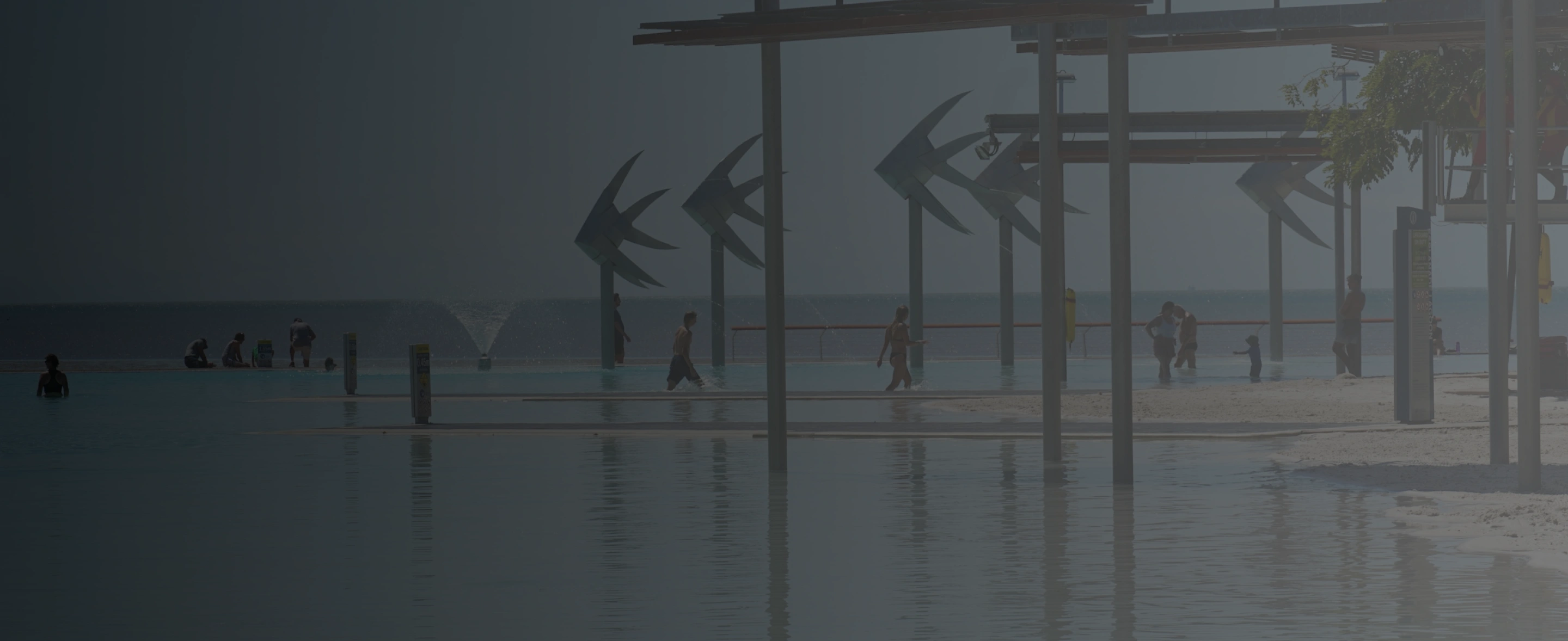 People walking and standing in a large outdoor pool with fish-shaped sculptures and a view of the ocean in the background.