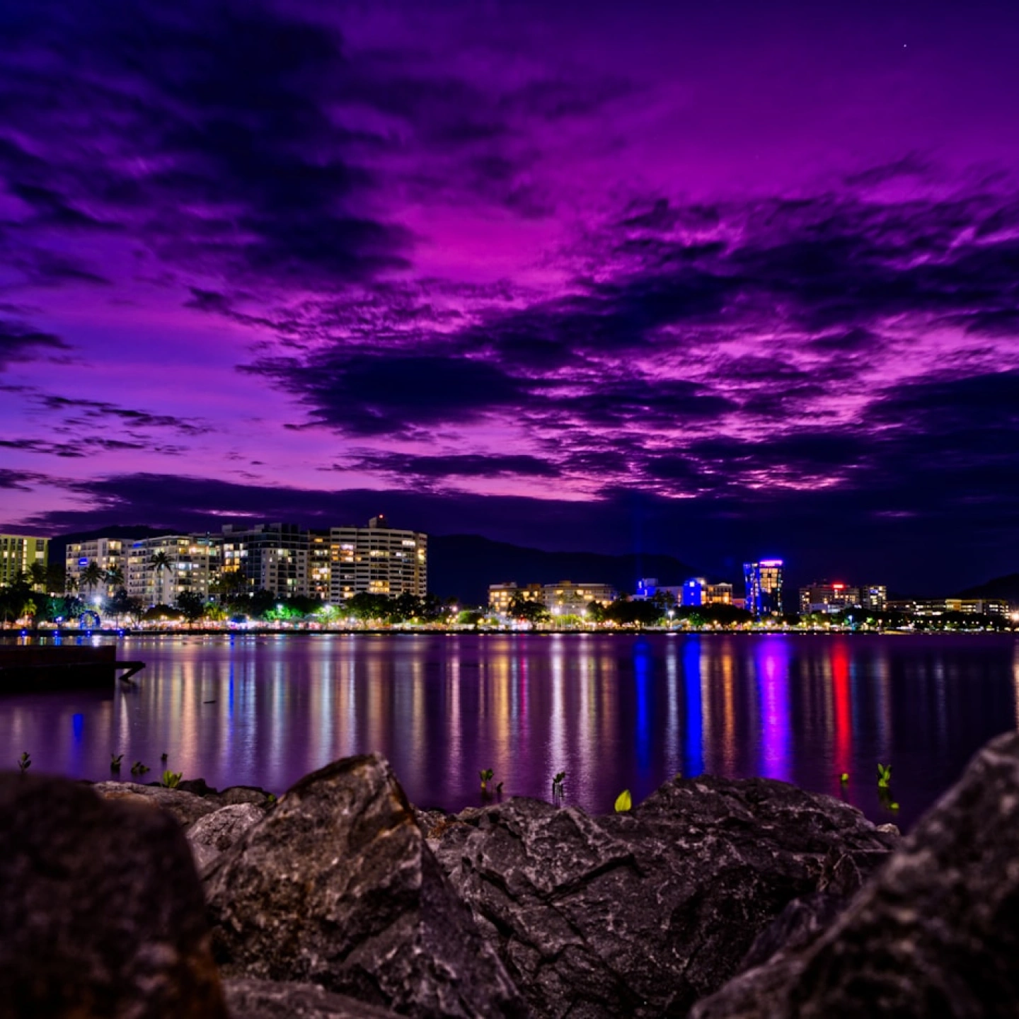 Night city skyline with illuminated buildings reflecting colorful lights on calm water under a vibrant purple sky.