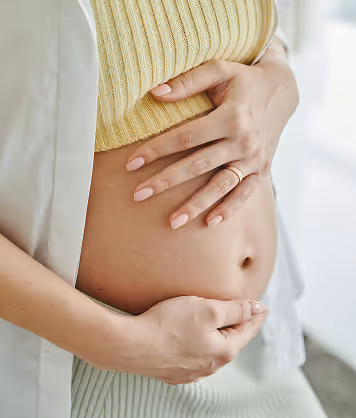 Close-up of a pregnant woman gently holding her bare belly with both hands, wearing a yellow knit top and a ring on her finger.