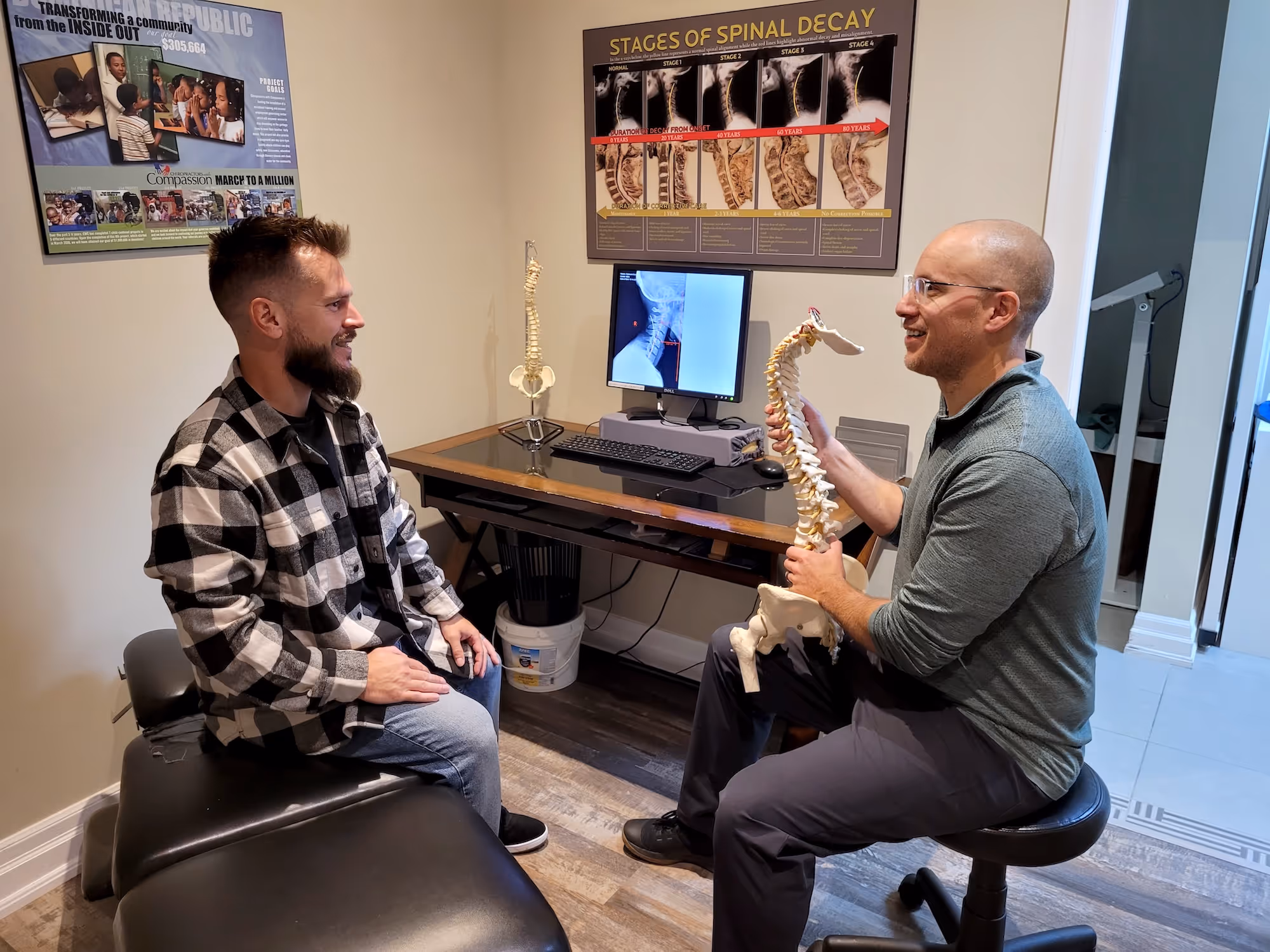Chiropractor explaining spinal anatomy to a patient using a spine model in an office with spine health posters.