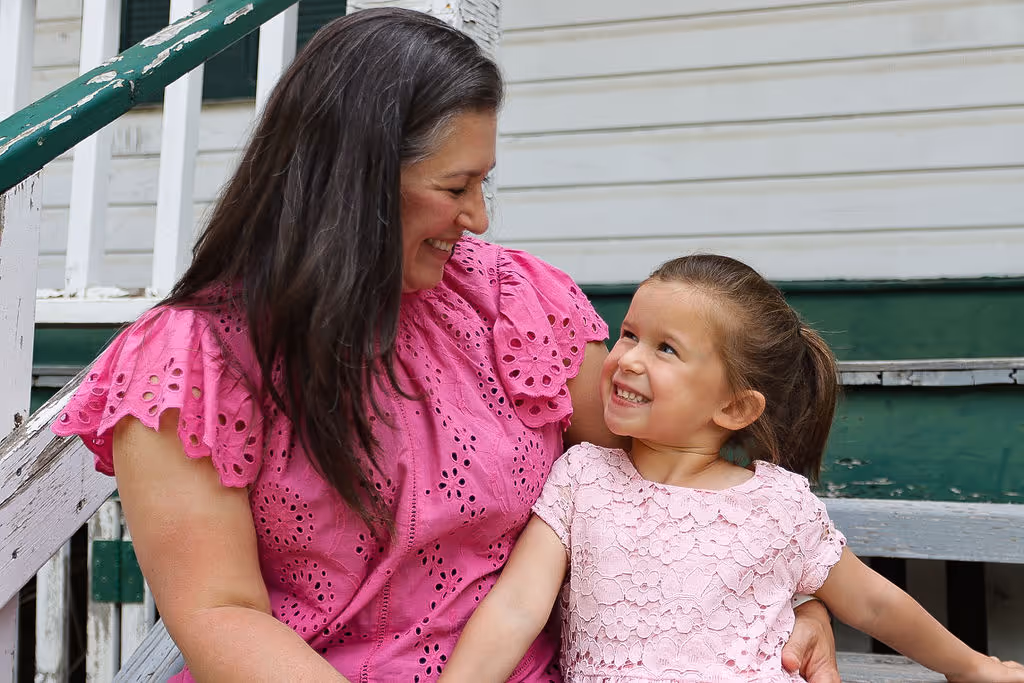 Woman in a pink eyelet blouse smiling warmly at a young girl in a pink lace dress sitting on her lap on wooden steps.