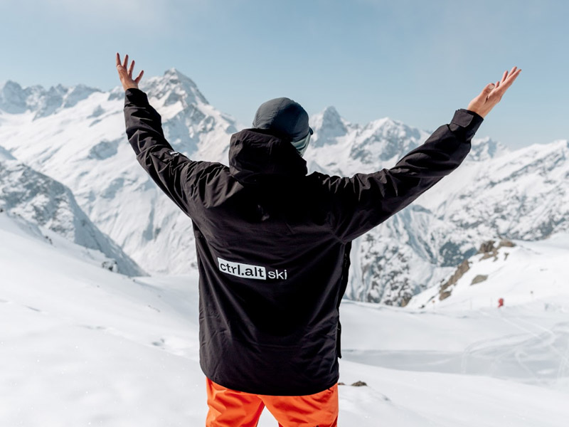A skier wears a branded ctrl.alt ski ski jacket, with his back turned to the camera and his hands in the air. He's surrounded by snowy mountainous peaks in the alps