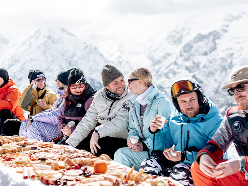 A group of ctrl.alt ski attendees enjoying a feast-like lunch on top of a mountain in the alps, two pictured are leaning into each other whilst the others are all laughing and smiling