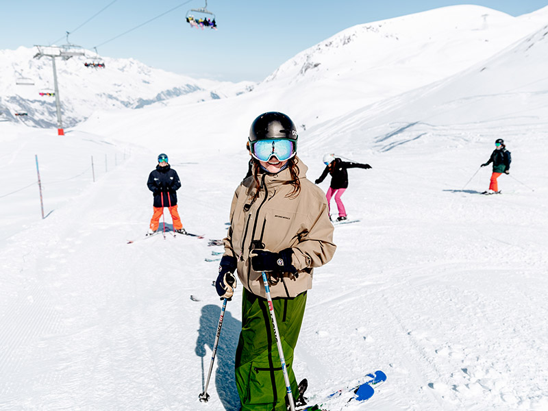 A skier stands in front of a mountainous backdrop with the sun reflecting from their blue goggles