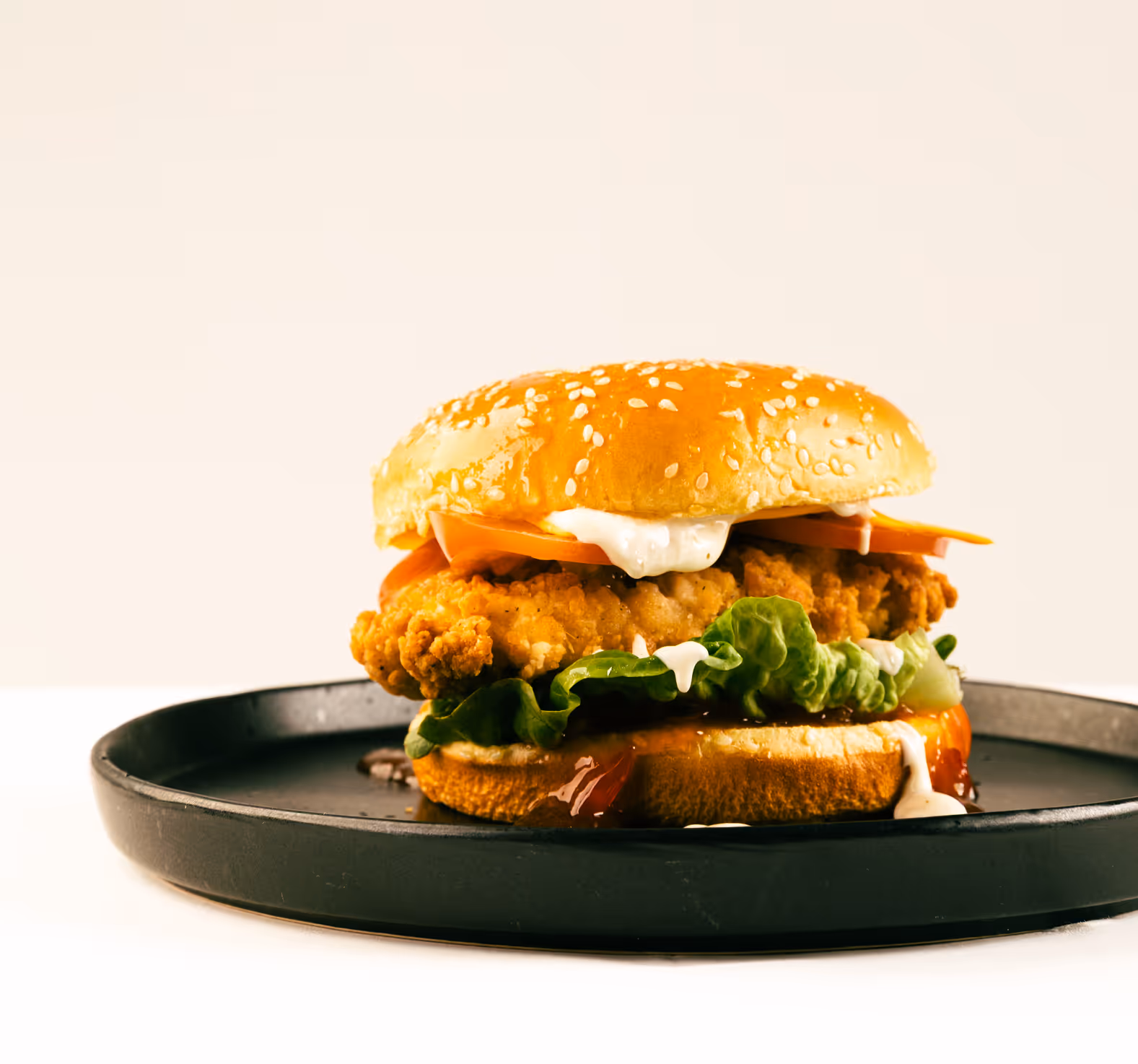 Crispy fried chicken sandwich with lettuce, tomato, and sauce on a sesame seed bun on a black plate.