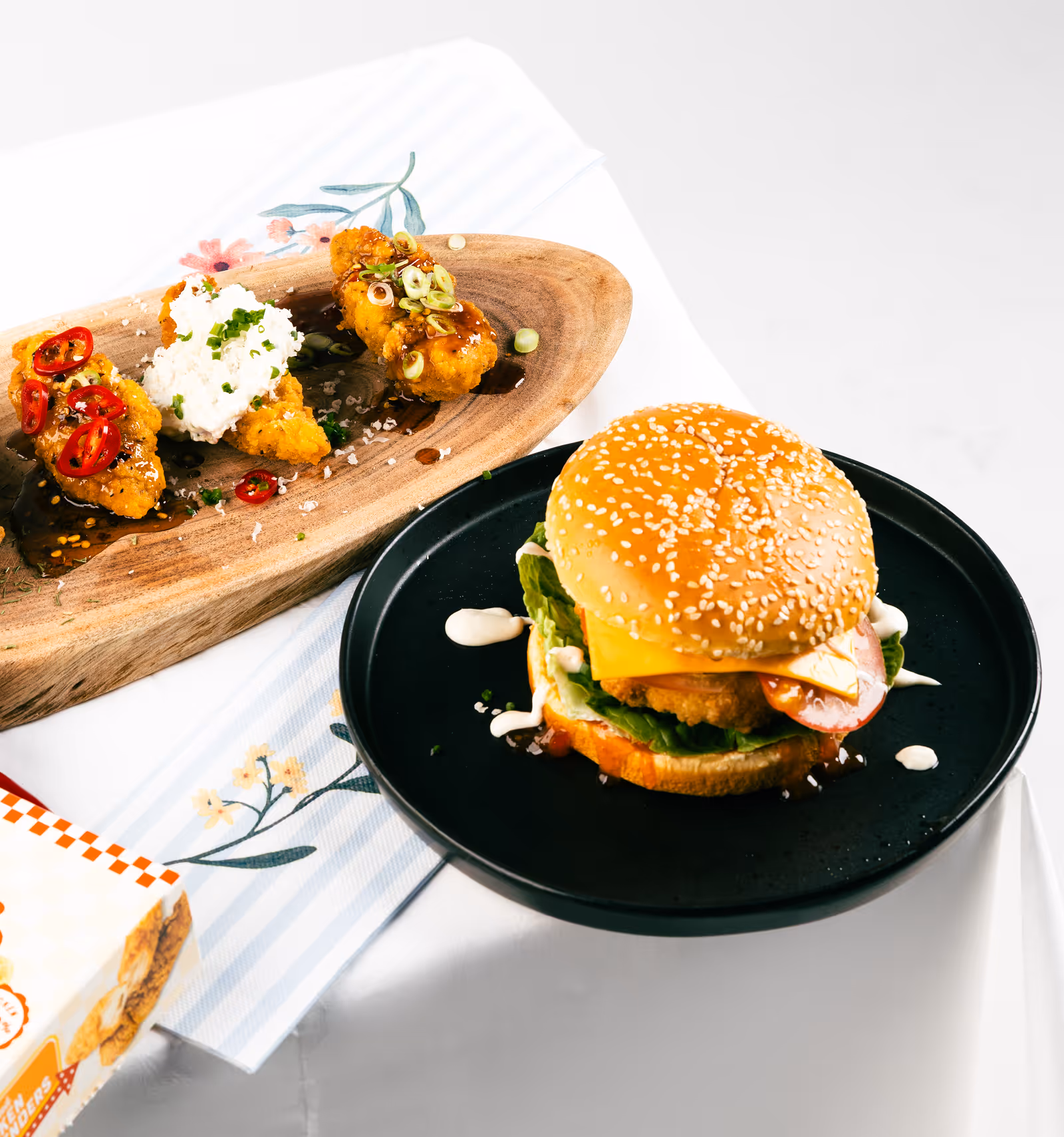 A crispy chicken sandwich with lettuce, cheese, tomato, and sauce on a sesame seed bun on a black plate, next to a wooden platter with three fried chicken tenders topped with sliced chili peppers, sour cream, and green onions.