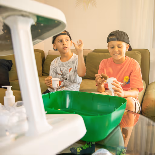 Two boys sitting on a couch, playing with toys and holding containers, with a large green plastic tray in the foreground.