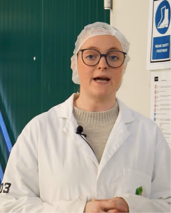 Person wearing a white lab coat, hairnet, and glasses standing indoors near a safety footwear sign.