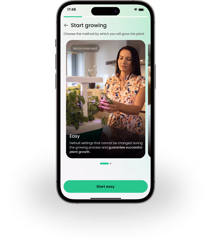 Mobile phone screen showing a plant-growing app with a smiling woman tending a hydroponic indoor garden under a lamp, labeled 'Easy' with recommended default settings for successful plant growth.