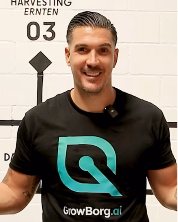 Man wearing a black GrowBorg.ai t-shirt explaining a green and white device on a table with bottles and other components.