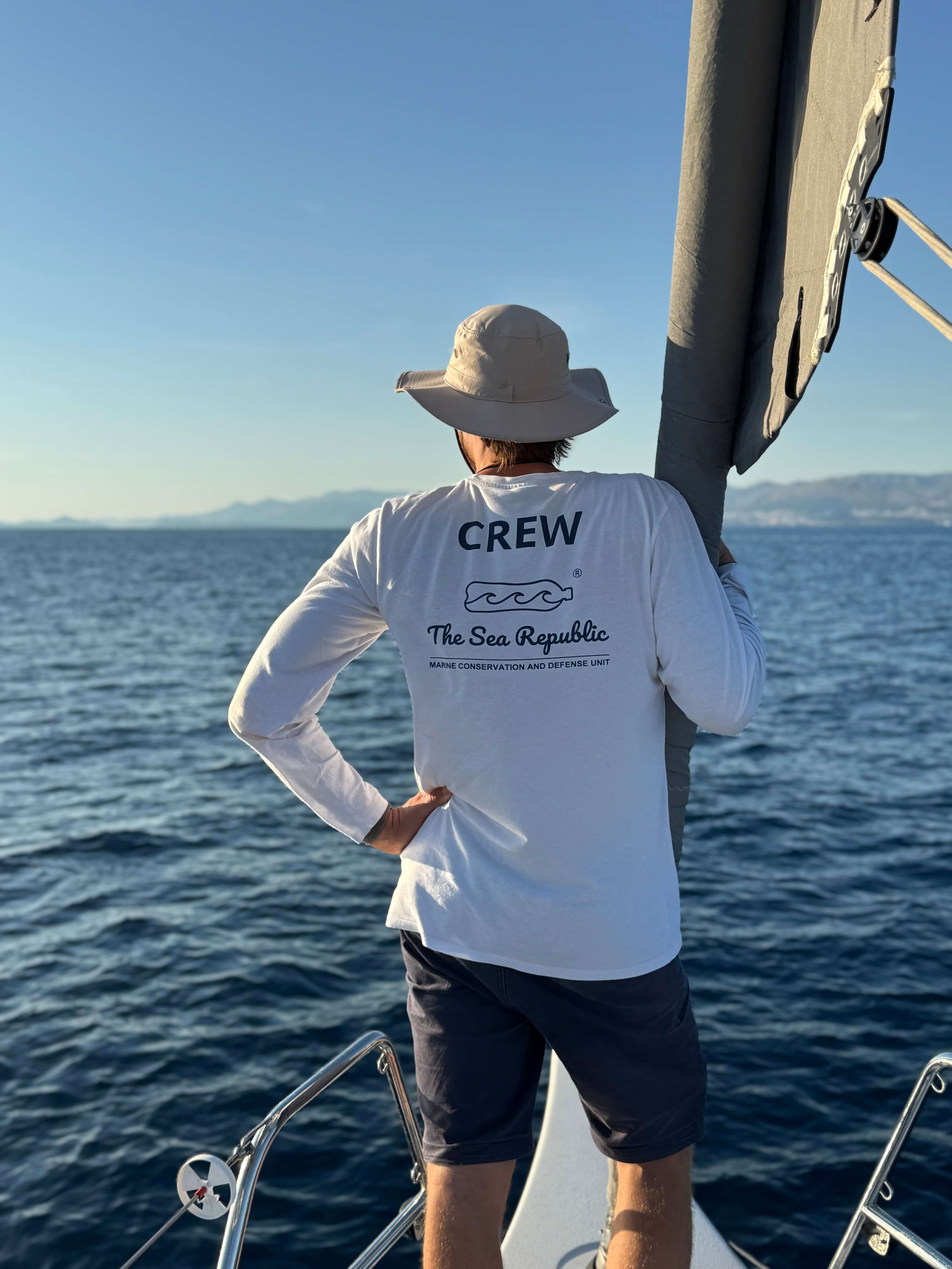 Persona in piedi su una barca che guarda il mare con maglietta bianca con scritta Crew sul retro.