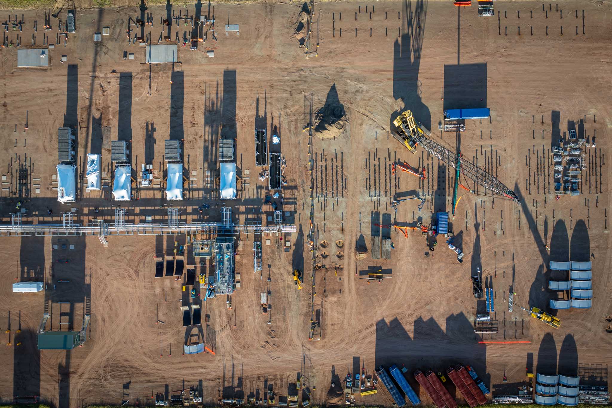 An aerial view looks straight down over a large industrial construction site with cranes, equipment, pipes and rows of structural posts. Long shadows across the dirt worksite show the scale and layout of the project from above.