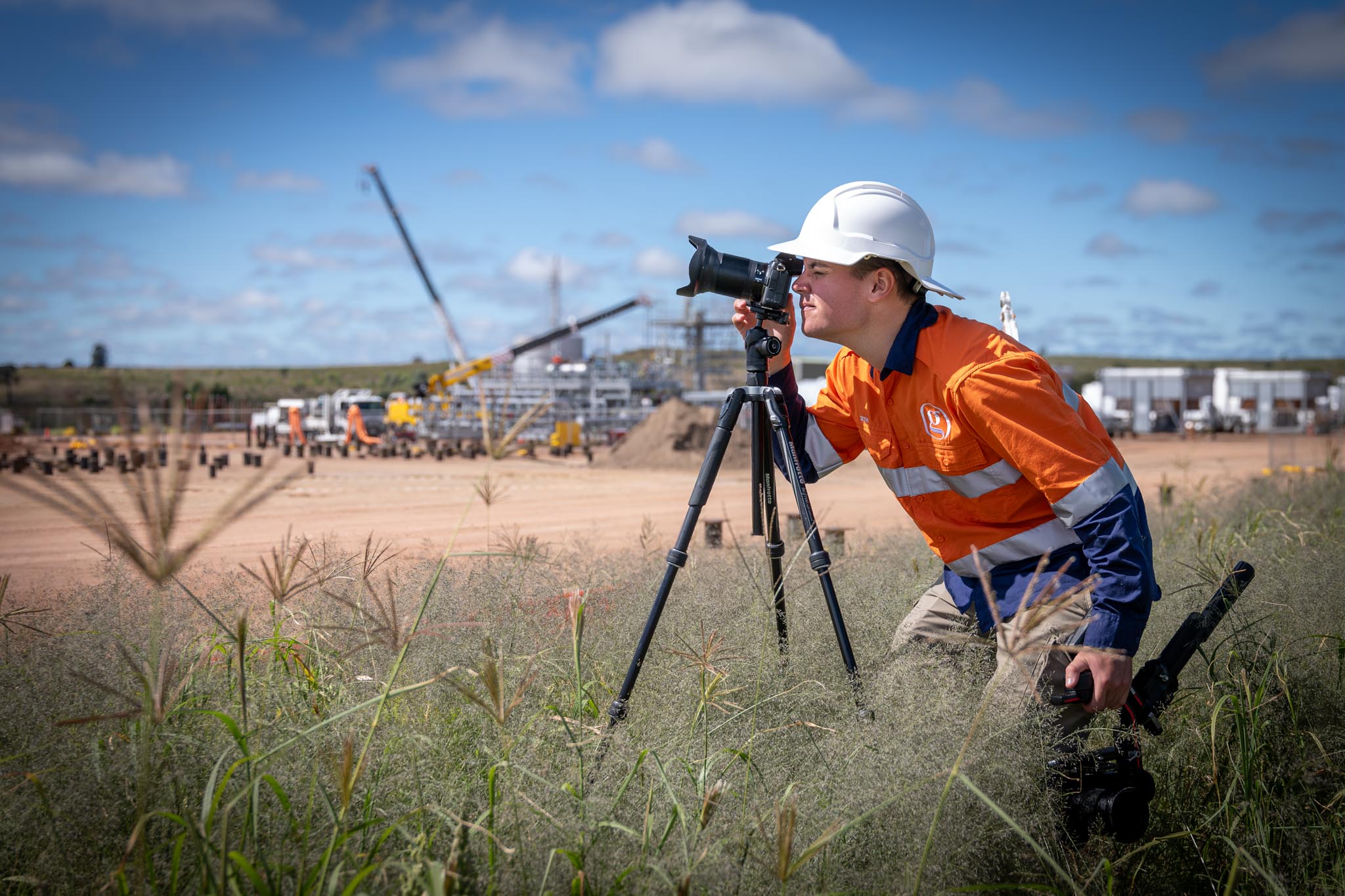 Seth Gerke from Gerkies looks through a camera on a tripod while crouching in grass near a construction site. Cranes, workers and site equipment in the background show him capturing professional industrial photography in the field.