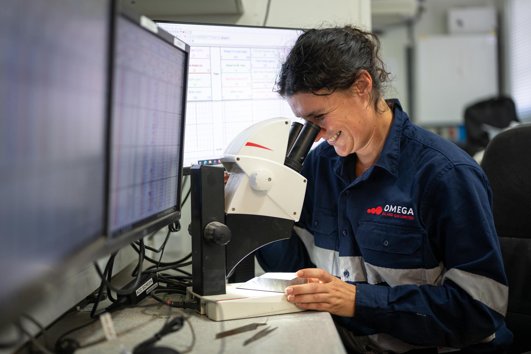 An Omega Oil and Gas worker smiles while looking through a microscope beside computer monitors in a lab workspace. The close up shows careful sample inspection or technical analysis in an oil and gas testing environment.