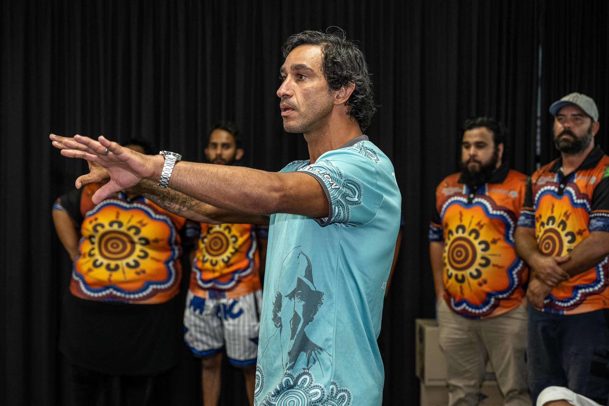 Jonathan Thurston gestures while speaking at an Indigenous Australian workshop or conference with several attendees standing behind him. The group wears colourful Indigenous patterned shirts in front of black curtains.