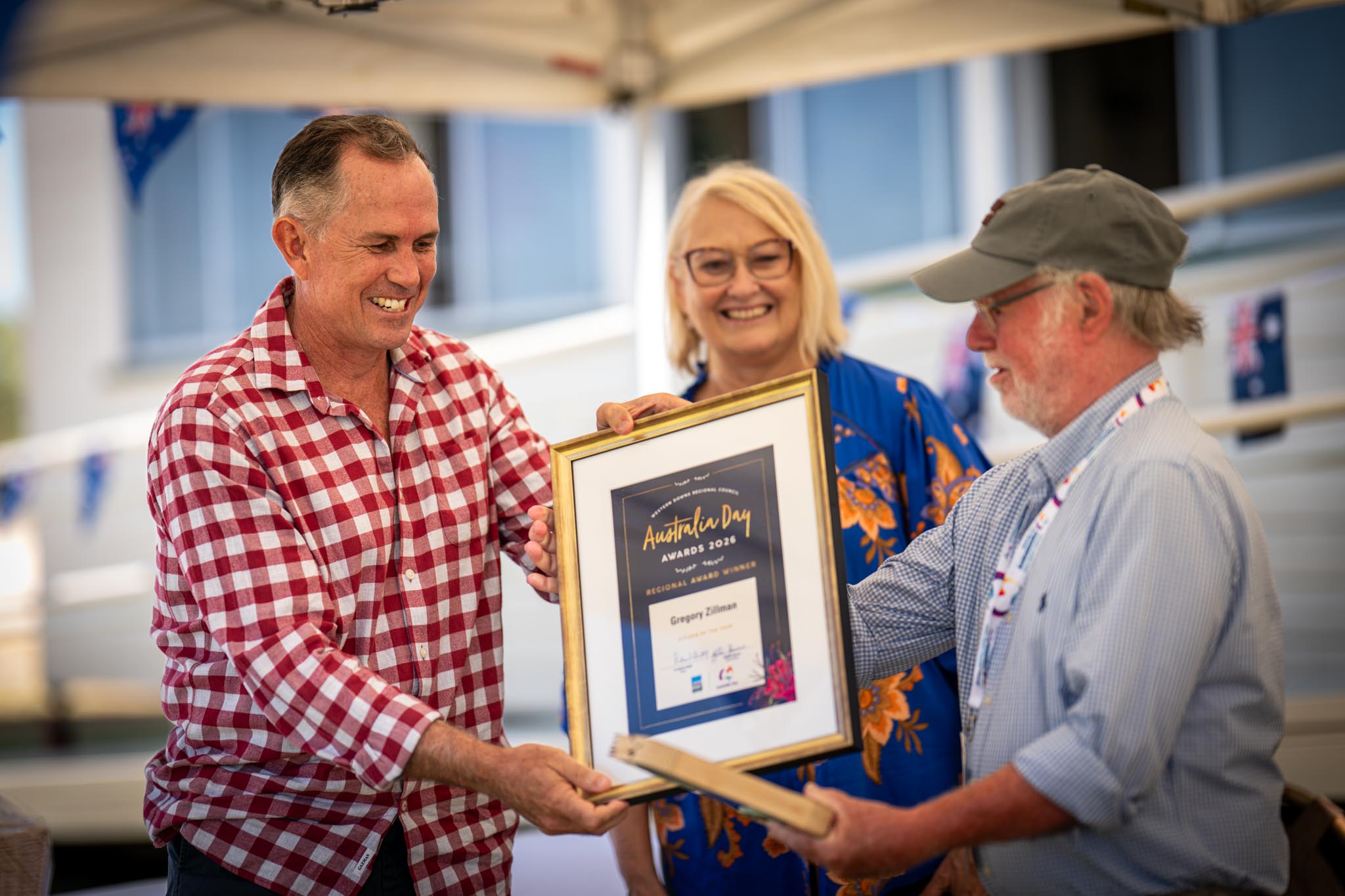 A Western Downs Australia Day Awards recipient smiles while receiving a framed certificate from two presenters at an outdoor ceremony. The certificate reads Australia Day Awards 2026 and the scene shows a formal community recognition moment.