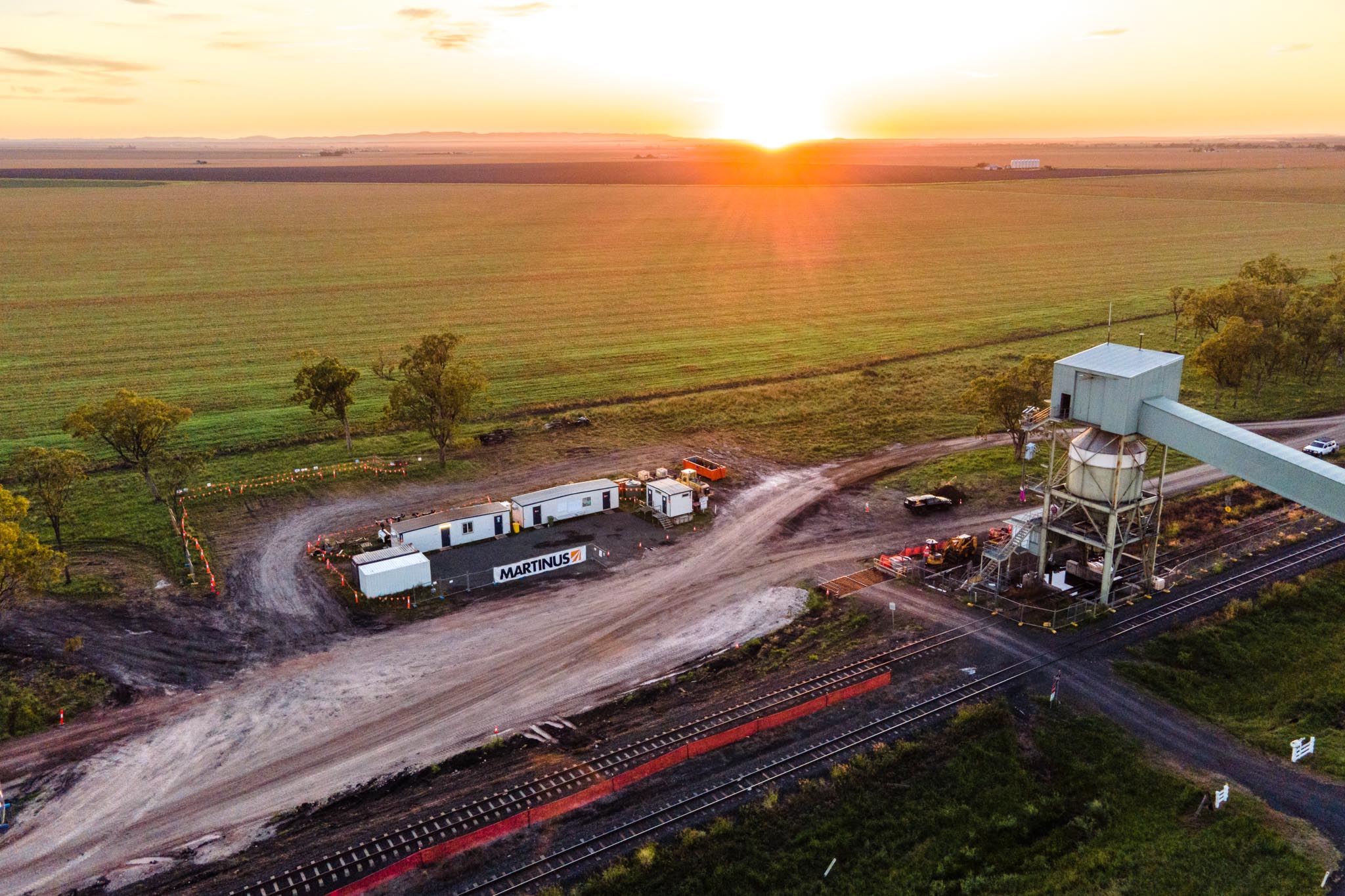 Martinus Rail site buildings and equipment sit beside railway tracks and a tall grain silo structure at sunset. The aerial view shows the rail worksite surrounded by open farmland and dirt access roads.