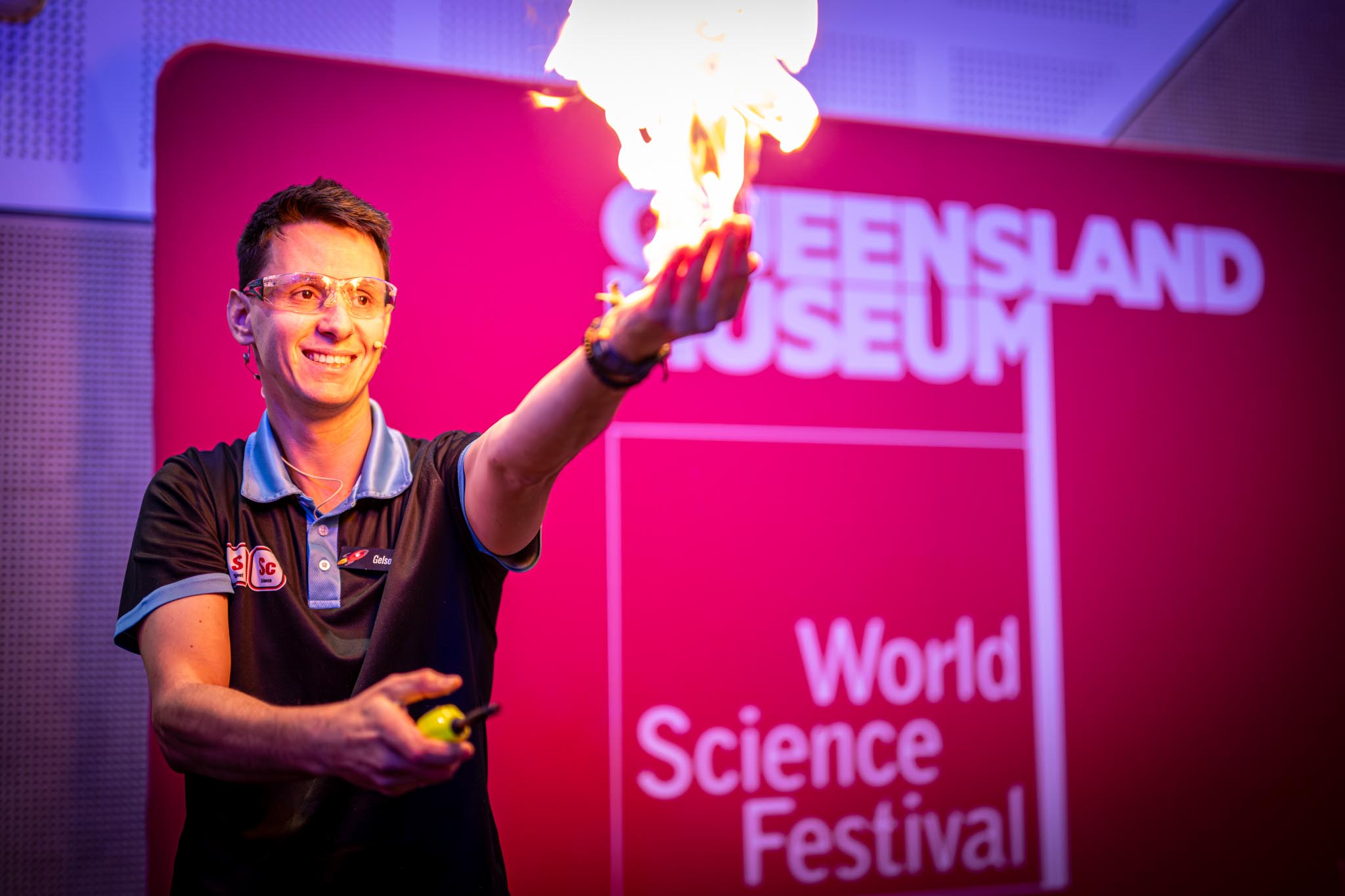 A science presenter holds a flame during a live demonstration at the World Science Festival by Queensland Museum. The background sign reads QUEENSLAND MUSEUM and World Science Festival against bright pink stage lighting.