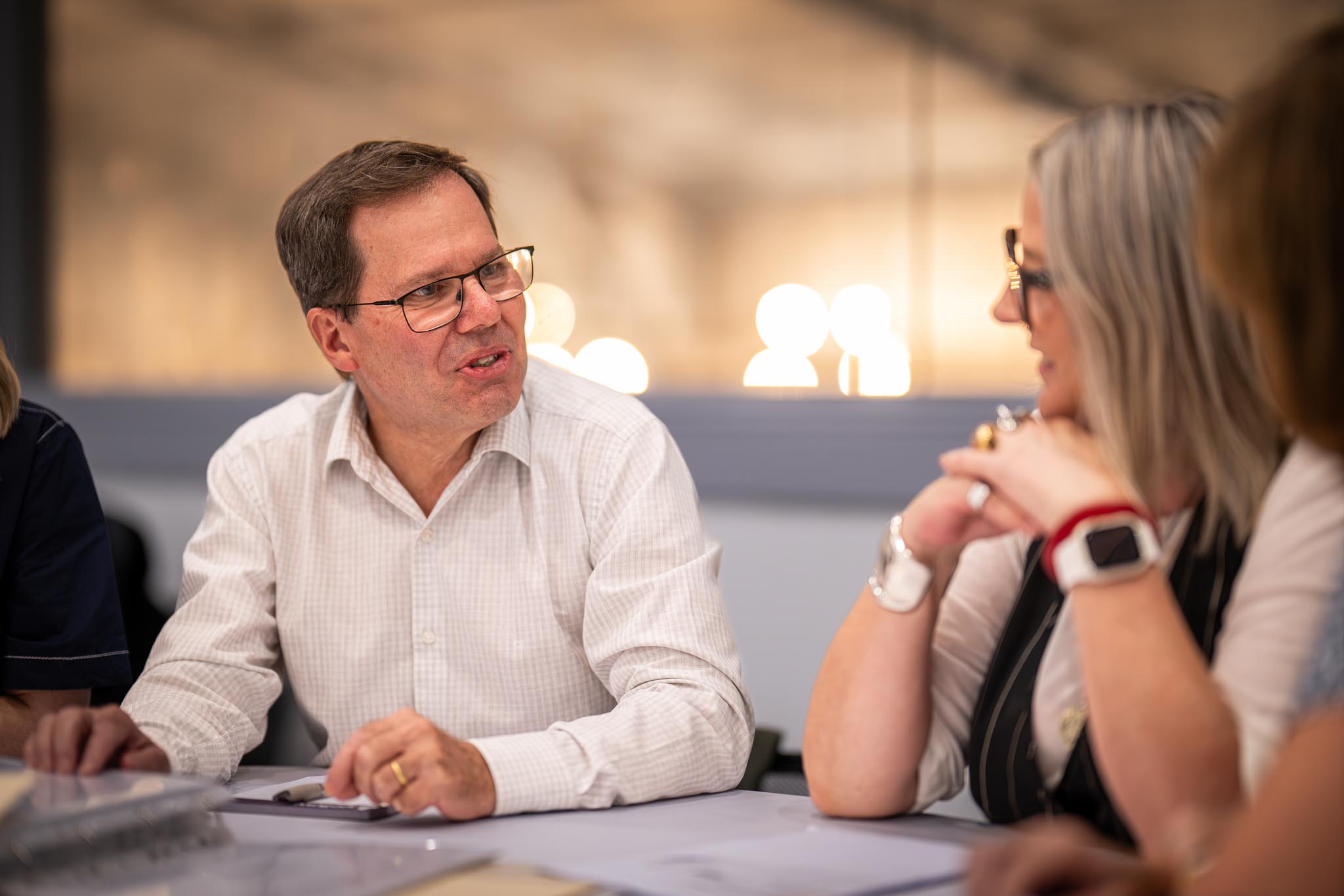 A man in glasses speaks with coworkers during a close up office meeting around a table. The warm lighting and attentive body language show a professional team conversation in progress.