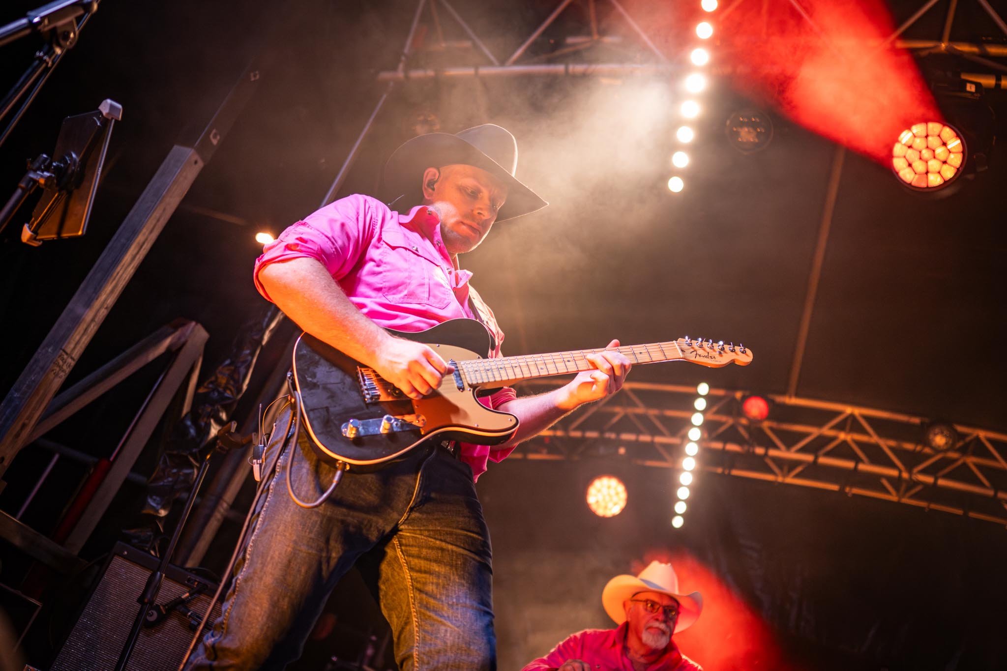 A guitarist in a cowboy hat and bright pink shirt performs on stage at Chinchilla Melon Festival. Red stage lights, smoke and another musician in the background create an energetic live music atmosphere.