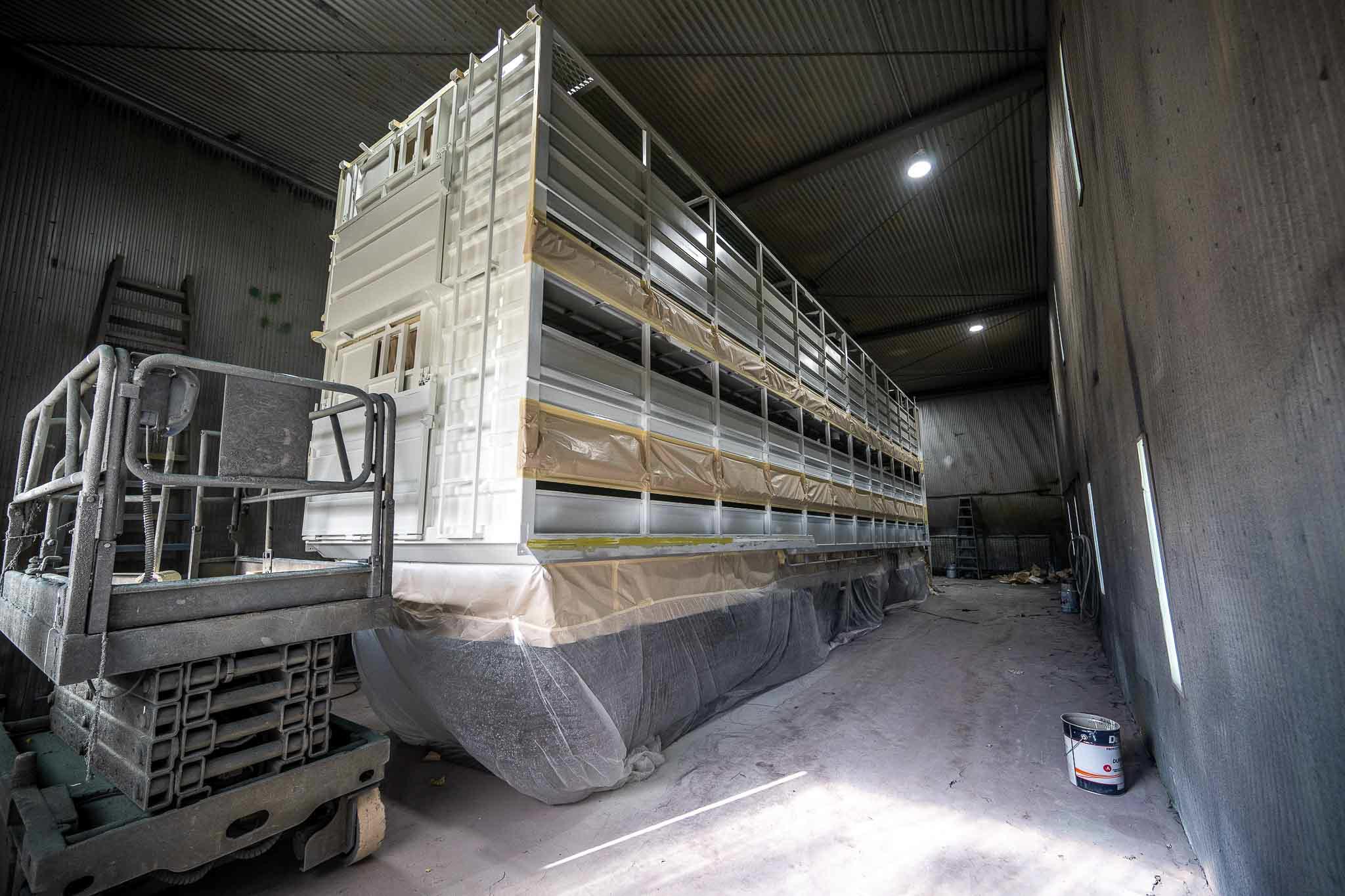 A large white industrial module sits inside a spray booth with masking tape and protective plastic covering sections of the structure. A scissor lift, paint bucket and bright wall lights show the scale of the coating or finishing work.