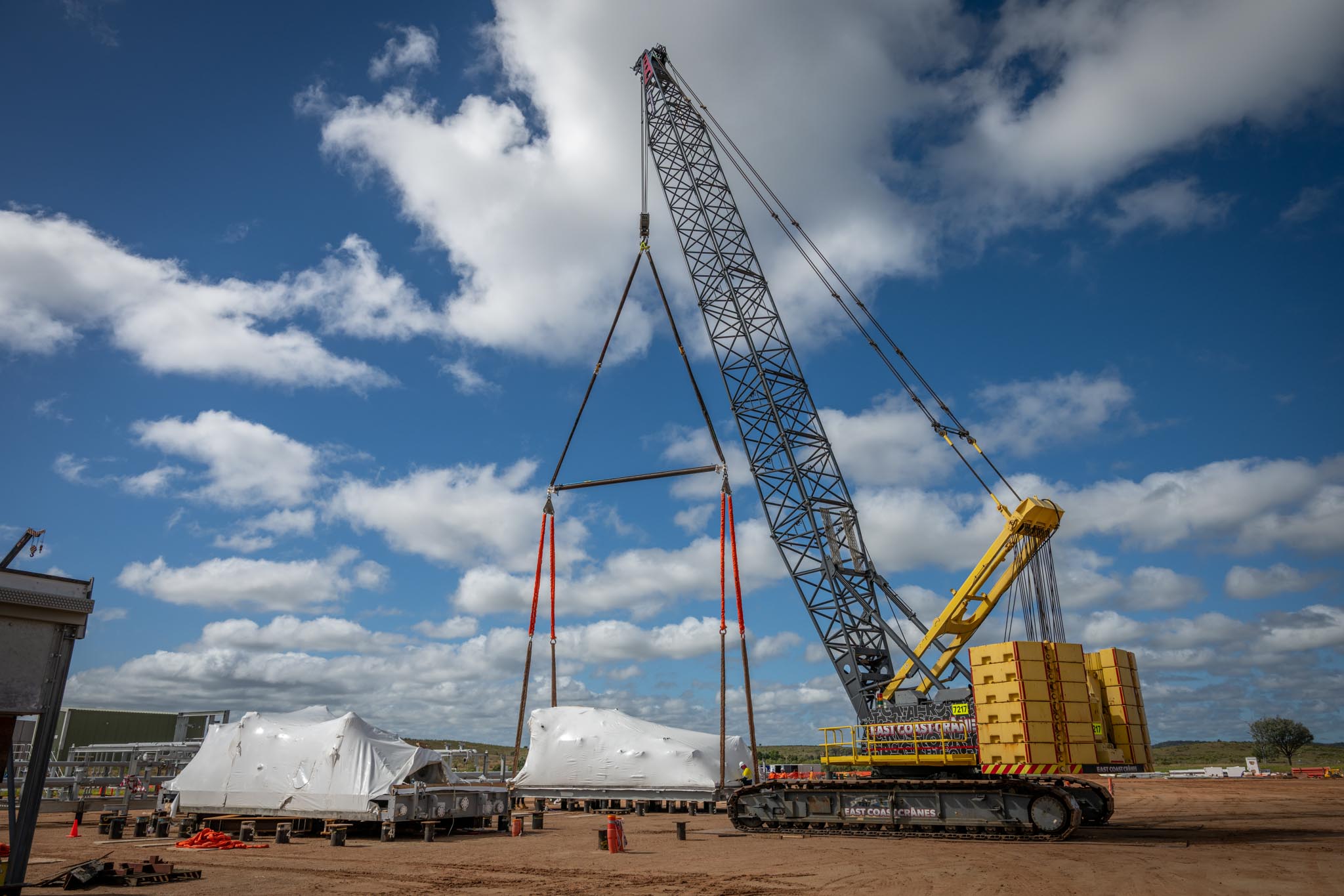 A large crawler crane lifts a wrapped industrial module at a construction site under a blue sky with scattered clouds. Heavy counterweights, lifting slings and another wrapped module nearby show a major industrial installation in progress.