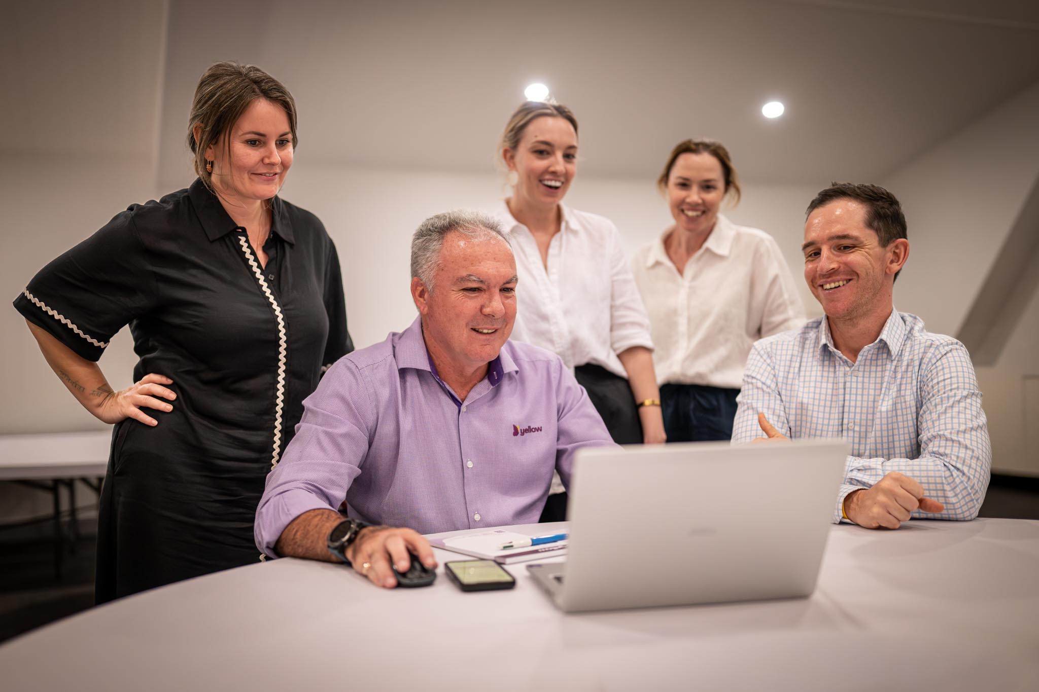 A group of five coworkers gather around a laptop during a relaxed office meeting. The team smiles while reviewing work together at a table with notebooks, pens and a phone nearby.