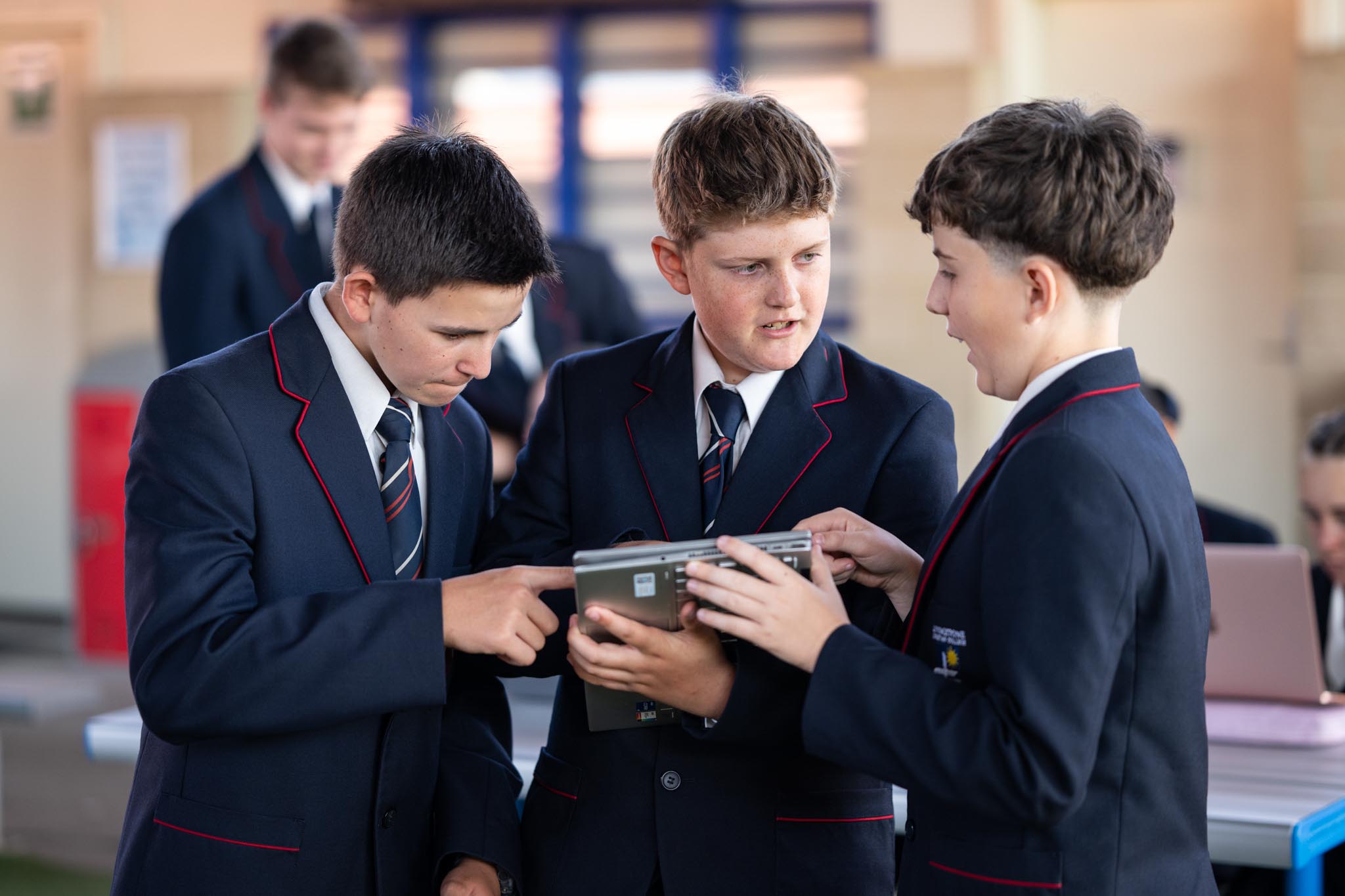 Three Livingstone Christian College students in school uniforms gather around a tablet during class. One student points at the screen while the others look closely, showing collaborative learning with technology.